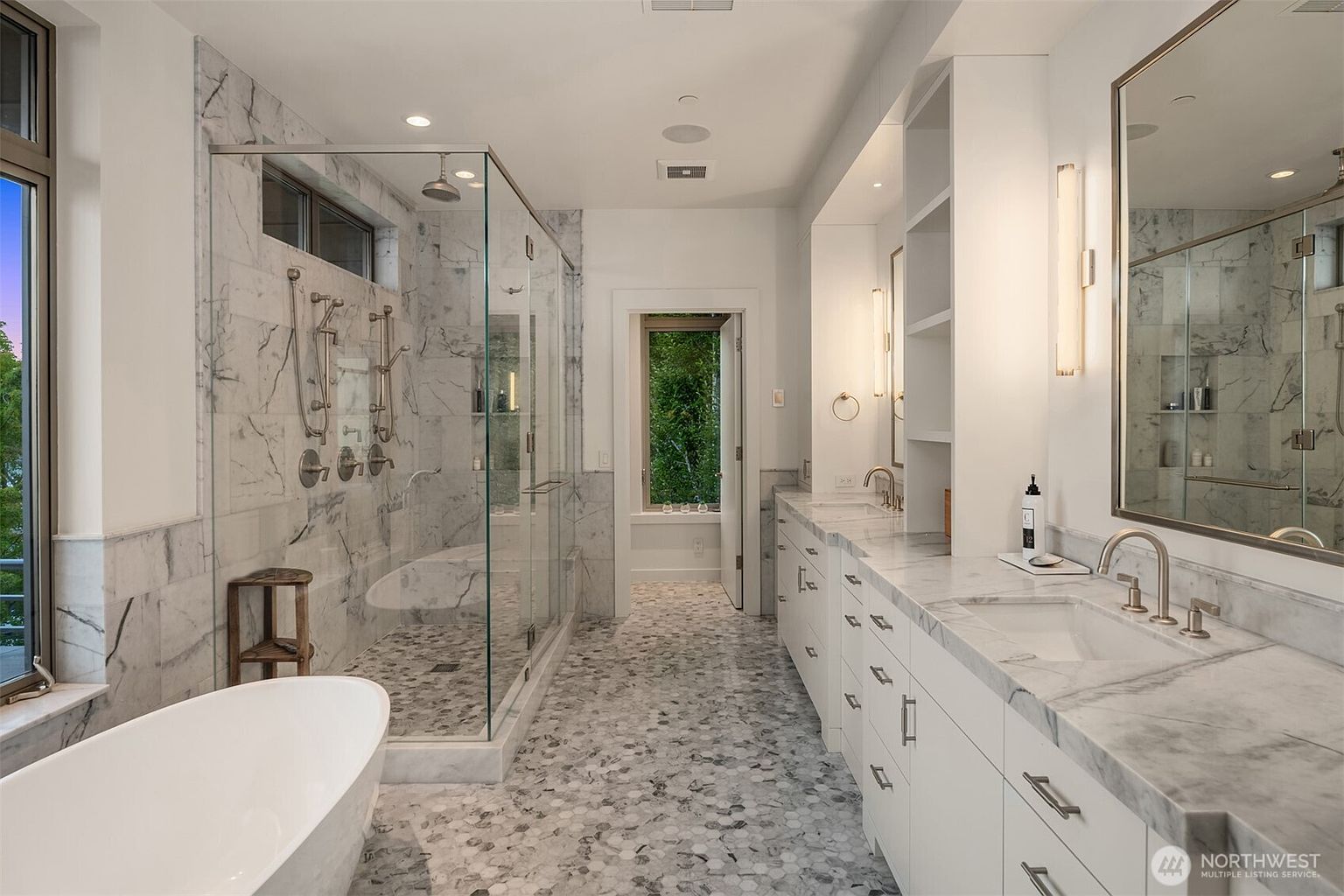 This luxurious primary bathroom features a sleek, modern design with a large glass-enclosed walk-in shower and a freestanding soaking tub. The space is finished with elegant marble tiling on the walls and a unique hexagonal mosaic floor, complemented by a long double vanity with marble countertops. A bright, airy atmosphere is created by the neutral color palette and the natural light streaming in from the window at the end of the room.