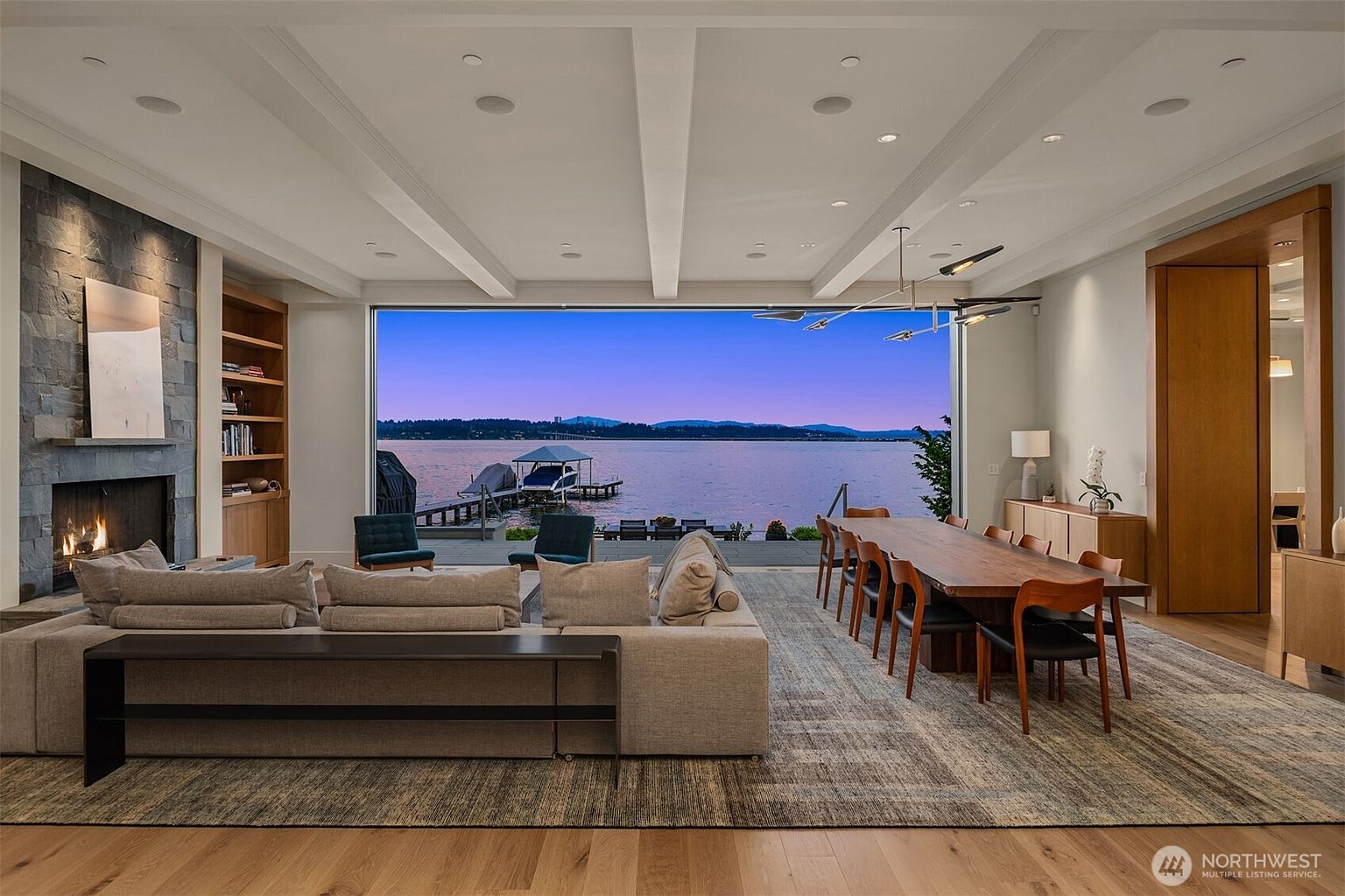 This expansive, open-concept living and dining area features a seamless transition to the outdoors through a massive floor-to-ceiling glass wall overlooking a serene lake. The space is anchored by a large, plush sectional sofa facing a striking stone fireplace, while a long, natural-edge wood dining table sits to the right, creating a sophisticated and airy atmosphere. The design emphasizes luxury and connection to nature, with high ceilings, recessed lighting, and a neutral color palette that highlights the stunning waterfront view.