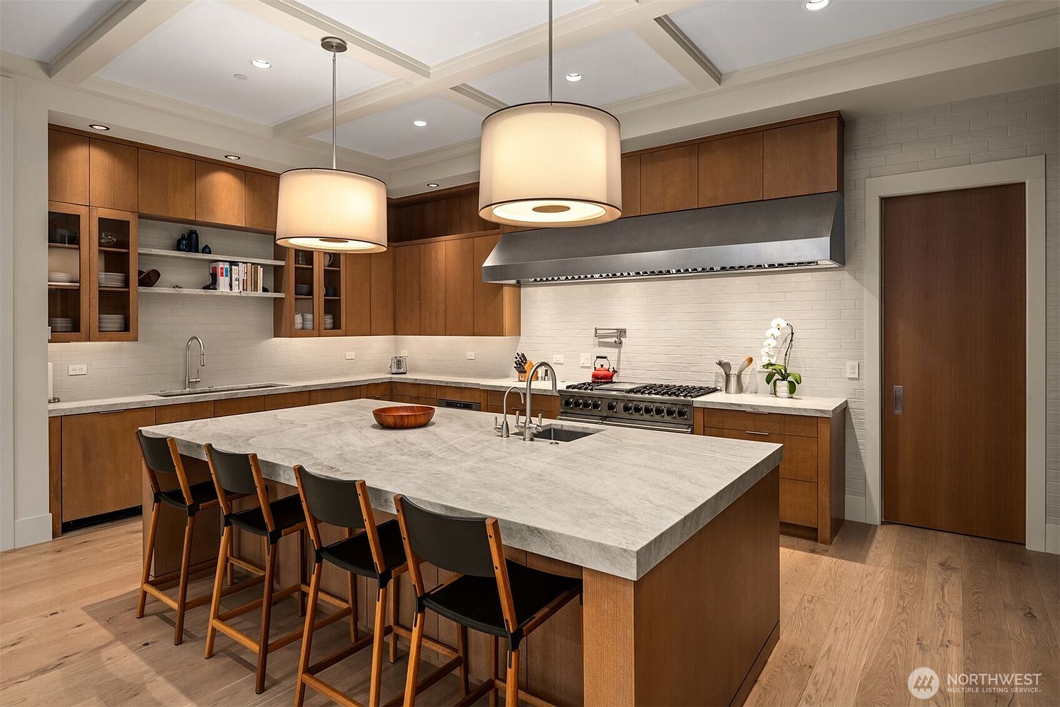 This sophisticated, modern kitchen features warm wood cabinetry, a large central island with a waterfall-edge stone countertop, and high-end stainless steel appliances. The space is illuminated by elegant drum pendant lights and recessed ceiling lighting, creating a bright and inviting atmosphere. A clean, minimalist aesthetic is complemented by a white subway tile backsplash and light-toned hardwood flooring, offering a perfect blend of functionality and luxury.