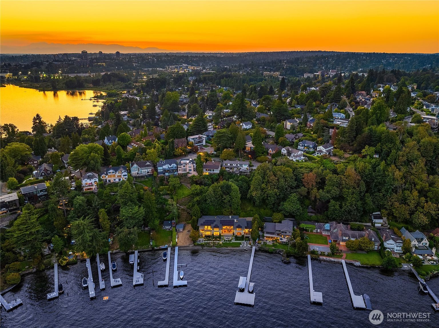 This stunning aerial view captures a luxurious waterfront property at sunset, showcasing its prime location along a serene lake. The perspective highlights the expansive shoreline, private docks, and the beautifully landscaped grounds surrounding the multi-level residence, all set against a vibrant, golden-hued horizon. The neighborhood is lush with mature trees, creating a peaceful and exclusive atmosphere perfect for high-end real estate.