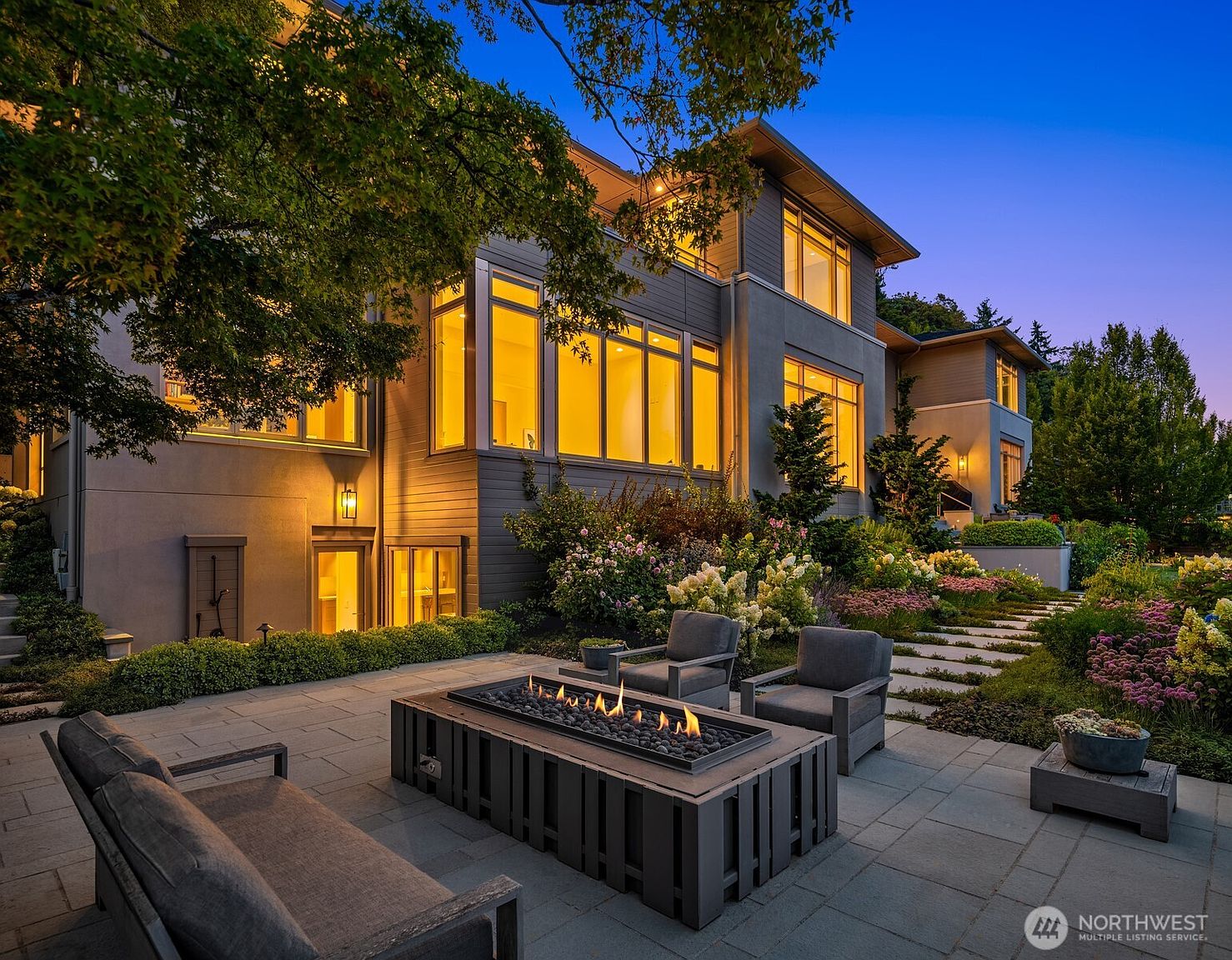 This elegant outdoor patio features a modern rectangular fire pit as the centerpiece, surrounded by comfortable lounge seating on a stone-paved terrace. The scene is set against the backdrop of a sophisticated, multi-story contemporary home with warm interior lighting glowing through large windows, creating an inviting and luxurious atmosphere. Lush landscaping, including flowering shrubs and a stone pathway, enhances the serene and upscale aesthetic of this backyard retreat.