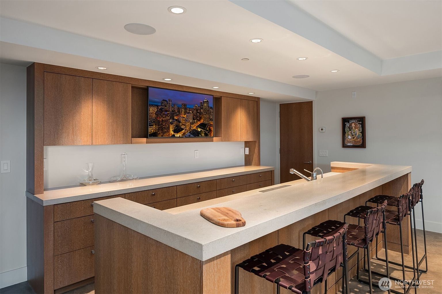 This modern, high-end wet bar or kitchen area features sleek, warm-toned wood cabinetry and a substantial concrete-style countertop. A wall-mounted television is integrated into the cabinetry, while a breakfast bar with woven leather stools provides a sophisticated seating area. The space is illuminated by recessed ceiling lights, creating a polished and inviting atmosphere perfect for entertaining.