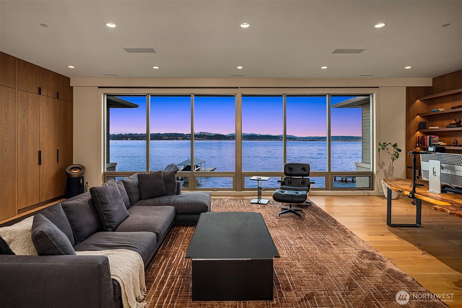 This sophisticated living room features a large, plush charcoal sectional sofa and a sleek black coffee table set atop a textured, earth-toned area rug. The space is anchored by a massive floor-to-ceiling window wall that offers a breathtaking, panoramic view of the lake at dusk. To the right, a live-edge wooden desk and a classic Eames-style lounge chair complete the room, blending modern luxury with a serene, natural backdrop.