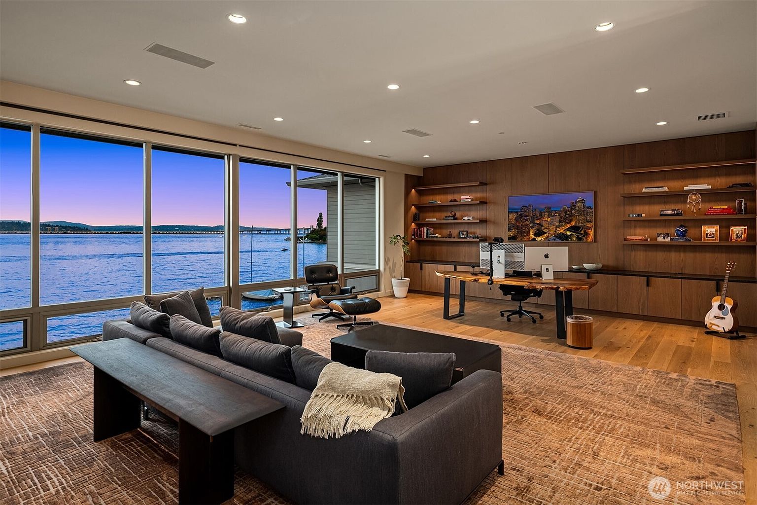 This sophisticated home office features a stunning live-edge wood desk positioned against a wall of warm, floor-to-ceiling wood paneling with integrated shelving and a television. A plush, dark gray sectional sofa sits on a textured area rug, facing a wall of expansive windows that provide a breathtaking, panoramic view of the water and distant shoreline. The room combines modern professional functionality with a serene, high-end aesthetic, perfect for both productivity and relaxation.
