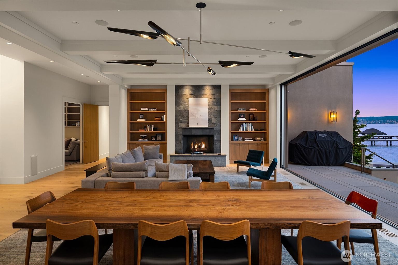 This expansive, open-concept living and dining area features a seamless transition to an outdoor deck overlooking the water through large sliding glass doors. The space is anchored by a striking stone fireplace flanked by custom wood shelving, complemented by a large, natural-edge wood dining table in the foreground. The modern aesthetic is enhanced by a sophisticated geometric chandelier and warm, neutral tones, creating an inviting and luxurious atmosphere.
