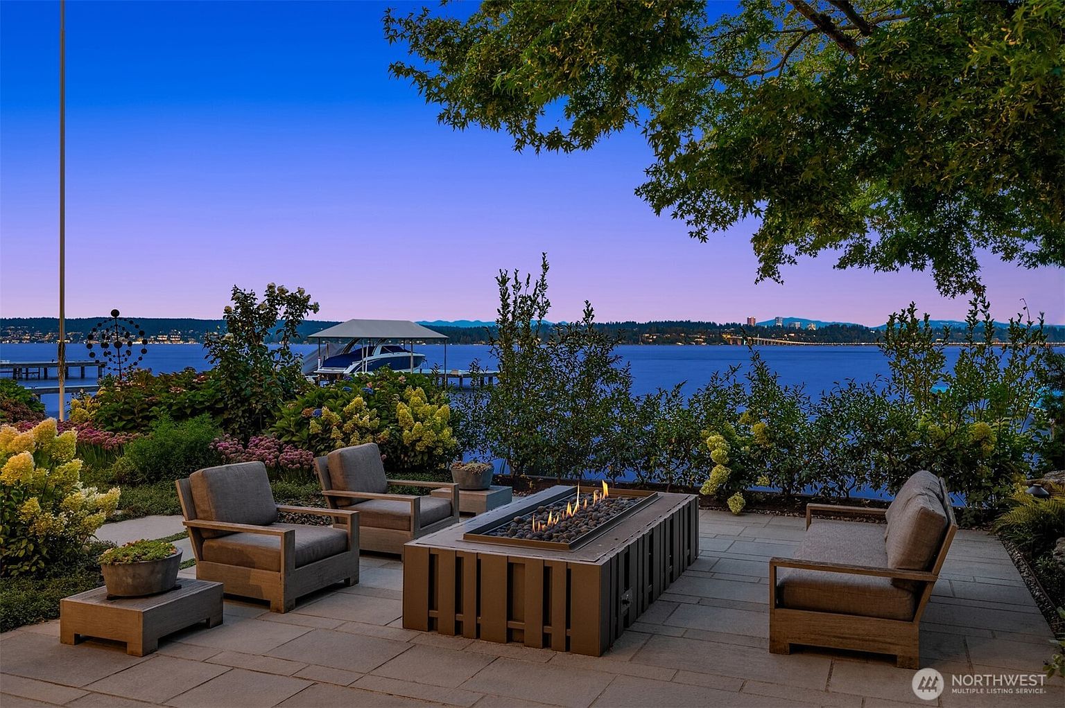 This stunning waterfront patio features a modern rectangular fire pit surrounded by comfortable, contemporary outdoor lounge furniture. The space is framed by lush landscaping and offers an expansive, unobstructed view of the lake and distant shoreline at dusk. The cinematic perspective captures a serene, high-end outdoor living area perfect for evening relaxation.