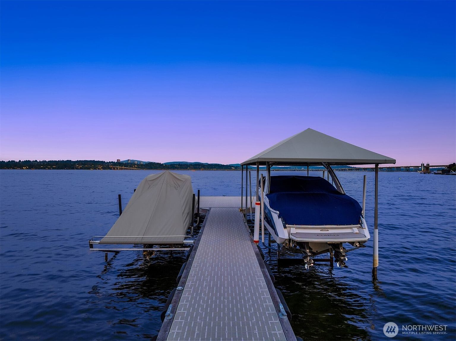 This image captures a serene, elevated perspective of a private boat dock extending into a calm lake at twilight. The dock features two boat slips, one covered with a canopy and housing a boat, and another with a boat under a protective cover. The horizon displays a soft gradient of blue and purple sky, creating a peaceful and luxurious waterfront atmosphere.
