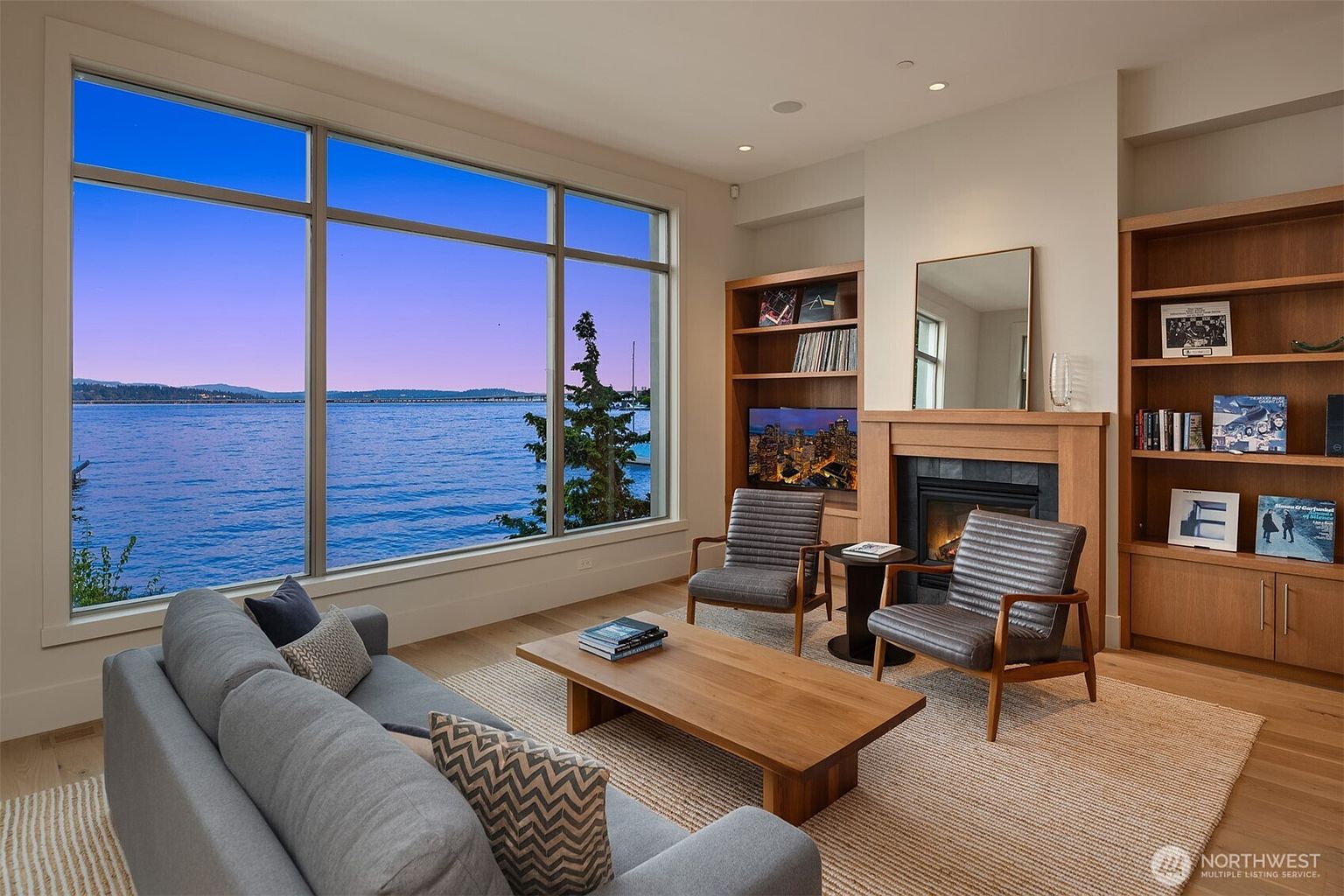 This sophisticated living room features a large, floor-to-ceiling window that frames a stunning, expansive view of the water and distant shoreline. The space is anchored by a cozy fireplace with built-in wooden shelving, two modern leather armchairs, and a plush grey sofa arranged around a minimalist wooden coffee table. The warm wood tones and neutral textures create a serene, high-end atmosphere perfect for relaxation while enjoying the scenic waterfront backdrop.