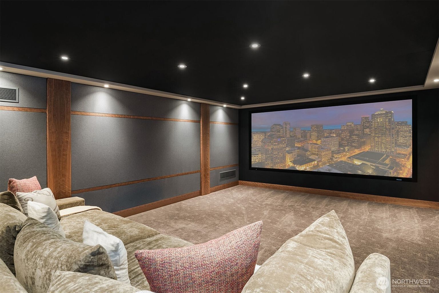 This image showcases a sophisticated, dedicated home theater room designed for an immersive cinematic experience. The space features plush, velvet-textured sectional seating, dark acoustic wall paneling with wood trim accents, and a large, high-definition projection screen displaying a cityscape. The room is finished with a dark ceiling and recessed lighting, creating a moody, professional-grade atmosphere perfect for entertainment.