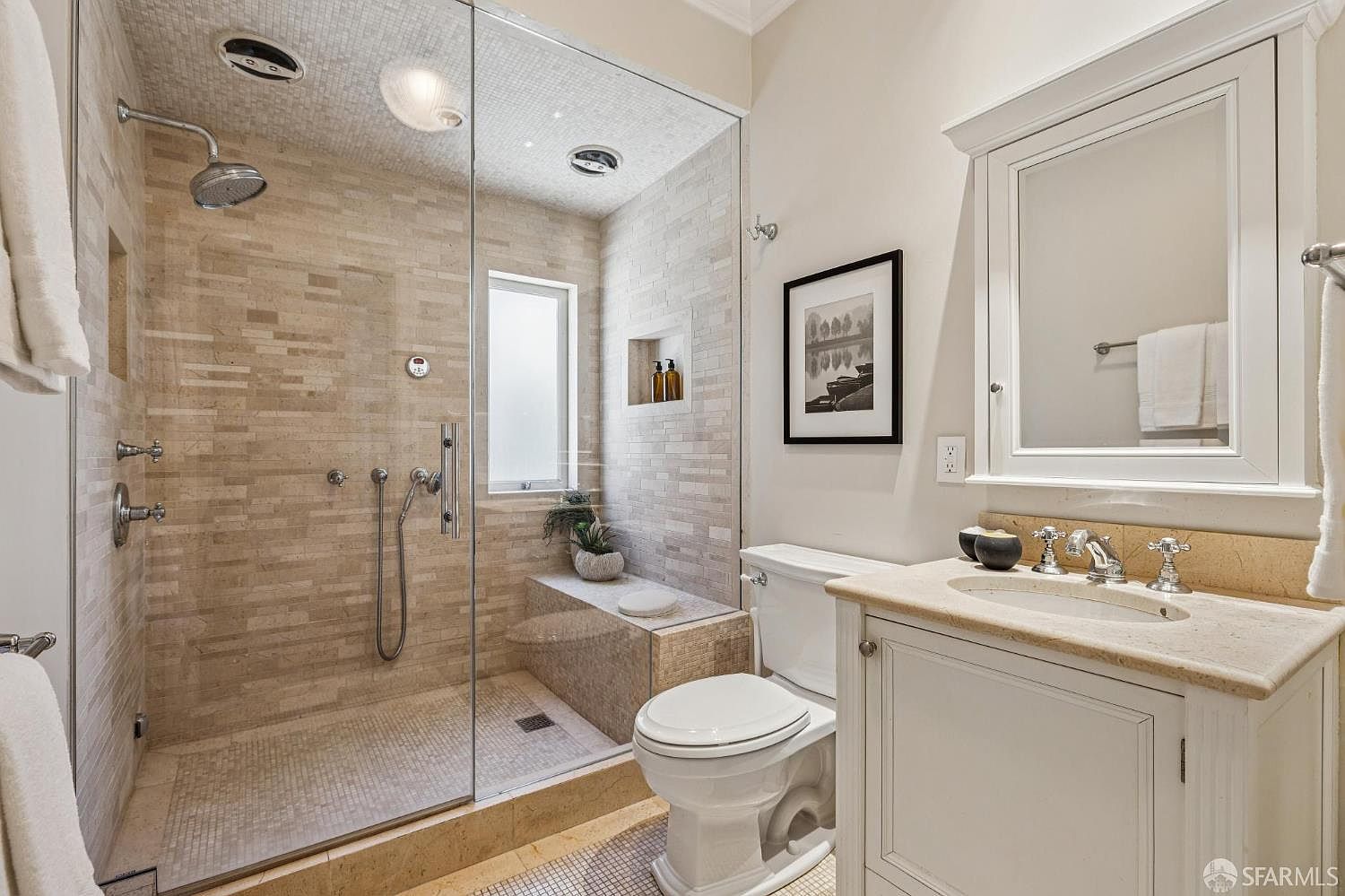 This compact, well-appointed bathroom features a walk-in shower with a glass enclosure, light-toned stone wall tiles, and a built-in bench. A white vanity with a stone countertop and a medicine cabinet mirror sits beside a standard toilet, creating a functional and cohesive layout. The neutral color palette and clean lines provide a bright, modern aesthetic suitable for a guest or secondary bathroom.