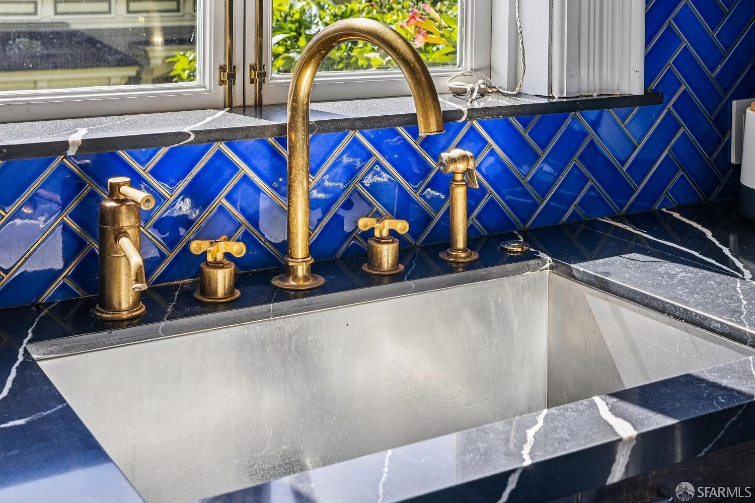 This close-up shot highlights a kitchen sink area featuring a metallic basin set into a dark, veined stone countertop. A prominent brass-toned faucet with matching handles and a side sprayer stands against a vibrant blue herringbone-patterned tile backsplash. The composition captures the elegant contrast between the rich blue tiles and the warm metallic fixtures, framed by a window that allows natural light to illuminate the workspace.