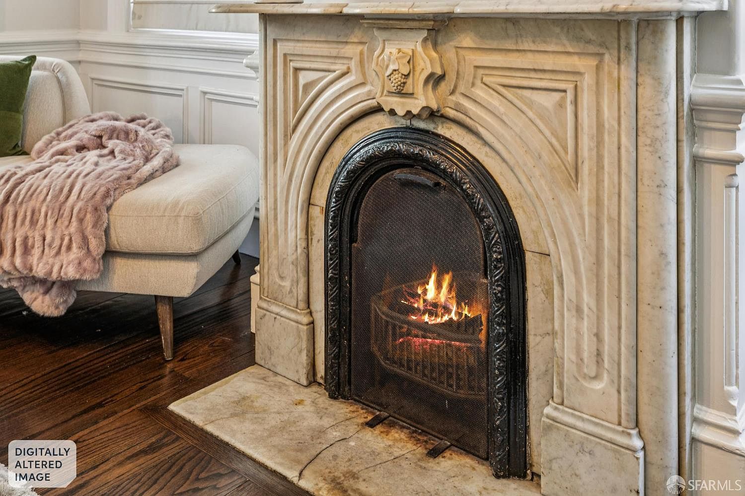 This interior view highlights a classic fireplace with an ornate, carved stone surround and a decorative metal fire screen. To the left, a portion of a light-colored upholstered chair is visible, draped with a soft, textured throw blanket, set against a backdrop of traditional wall paneling. The warm glow from the fire creates an inviting atmosphere, complemented by the rich, dark wood flooring.