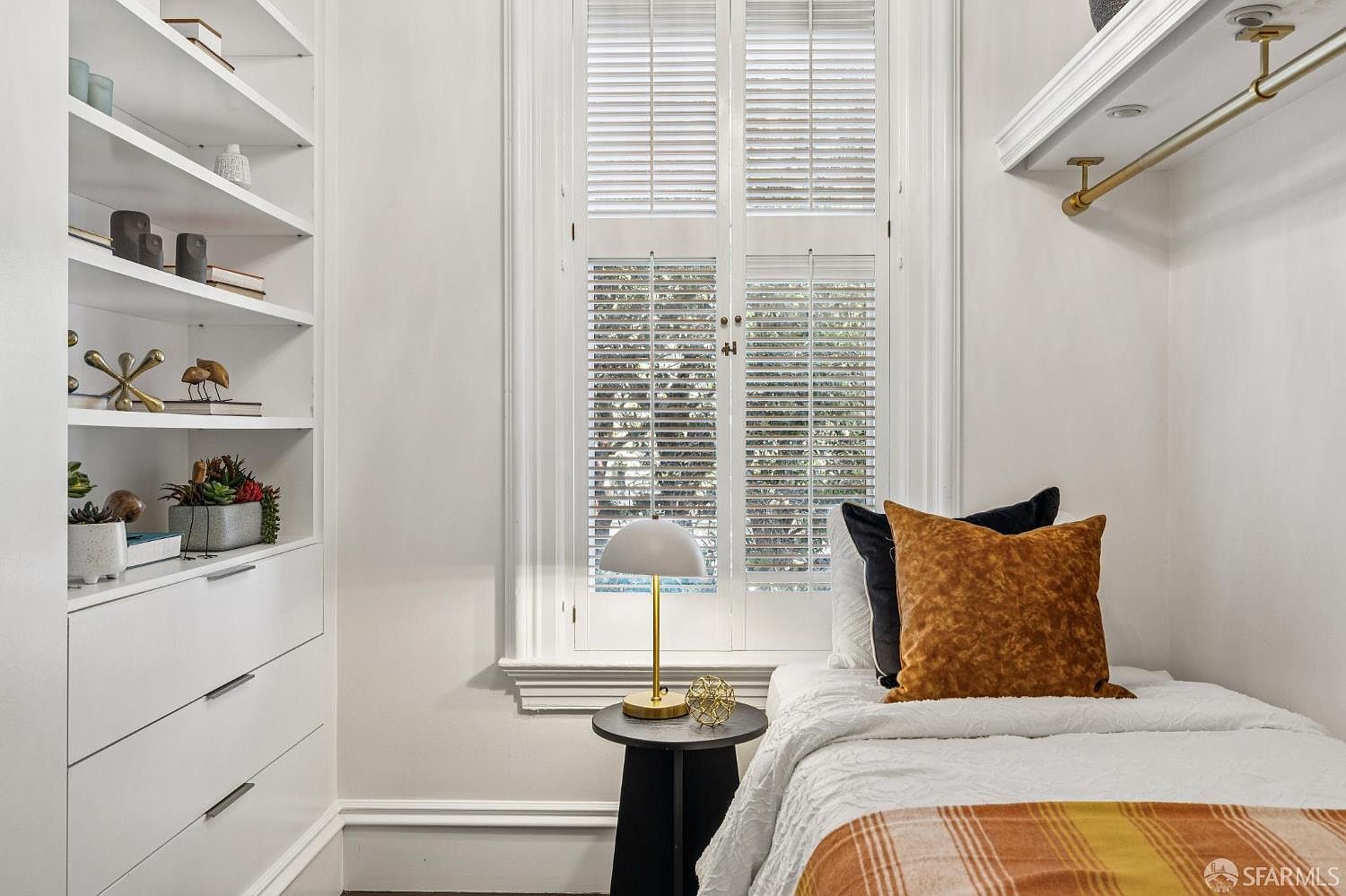 This cozy bedroom features a built-in shelving unit with drawers, providing ample storage alongside a twin bed dressed in neutral and warm-toned bedding. A window with white plantation shutters serves as the focal point, flanked by a small side table topped with a modern dome lamp. The room is bright and compact, offering a clean, organized aesthetic suitable for a guest space or home office nook.