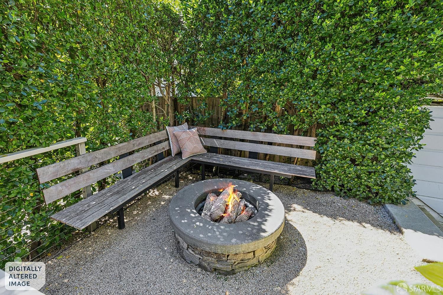 This cozy outdoor seating area features an L-shaped wooden bench nestled against a lush, green hedge, creating a private and intimate atmosphere. A circular stone fire pit with a burning fire serves as the focal point, set upon a gravel patio surface. The perspective is a slightly elevated, eye-level shot that captures the inviting, relaxed nature of this backyard retreat.