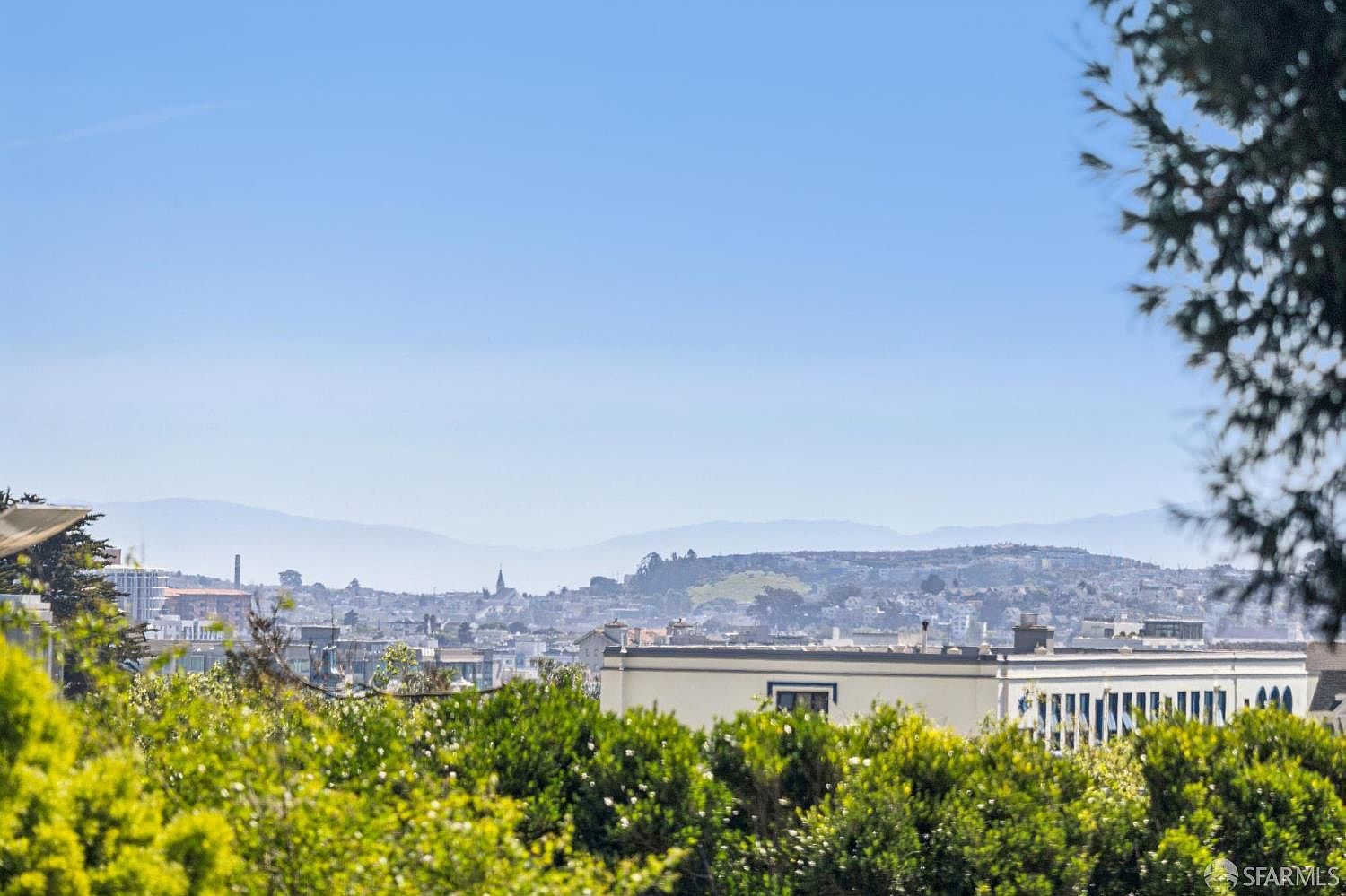 This image captures a scenic, elevated view of a cityscape from a lush, green vantage point. In the foreground, vibrant green foliage frames the scene, while the mid-ground features a flat-roofed building and a distant church steeple set against a sprawling urban landscape. The background showcases rolling hills under a clear, bright blue sky, creating a peaceful and expansive atmosphere.