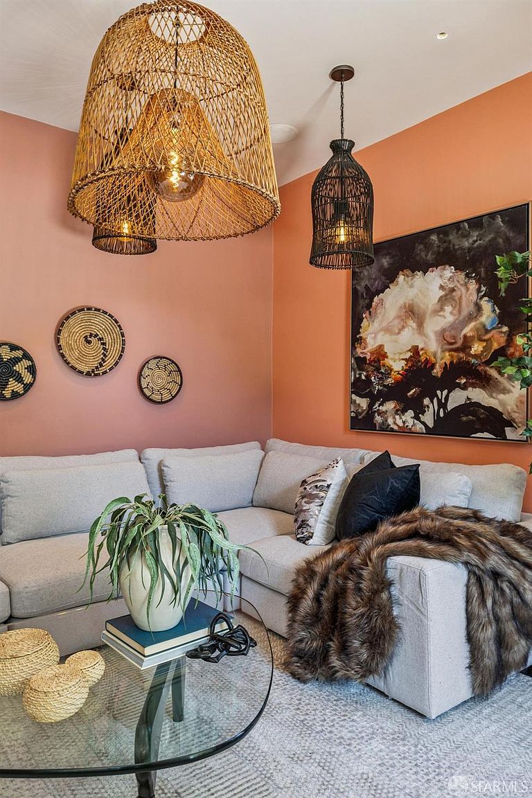 This cozy living room features a large, light-colored sectional sofa adorned with patterned and solid black throw pillows and a plush faux-fur throw. The space is accented by a warm, peach-toned wall, a large abstract art piece, and two distinct woven pendant lights hanging above. A glass-topped coffee table holds a potted plant and decorative woven baskets, creating a stylish and inviting atmosphere.