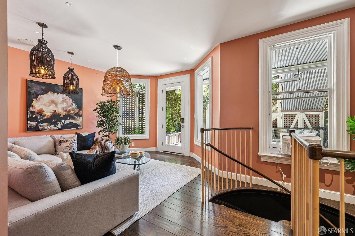 This inviting living room features warm, peach-toned walls and rich dark wood flooring, creating a cozy and vibrant atmosphere. The space is furnished with a plush, light-colored sectional sofa adorned with decorative pillows, a glass-topped coffee table, and a large abstract art piece. A spiral staircase with a wooden railing leads to a lower level, while multiple windows and a glass door provide ample natural light and a view of the outdoor greenery.