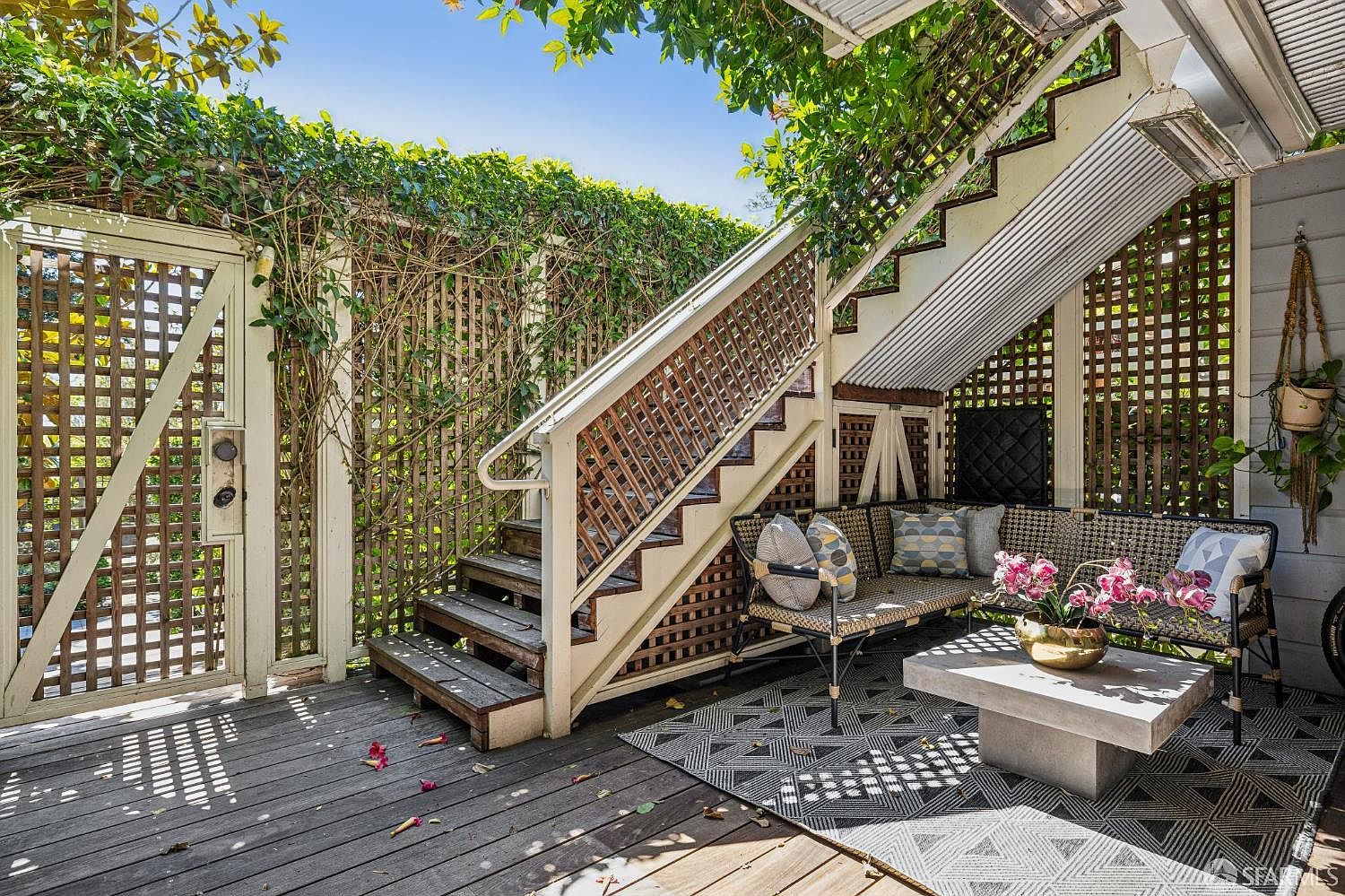 This inviting outdoor deck features a cozy seating area tucked beneath a staircase, complete with a woven sofa, patterned cushions, and a modern concrete coffee table. The space is framed by decorative wooden lattice walls covered in lush climbing vines, creating a private and serene atmosphere. A geometric patterned rug anchors the seating area, while the wooden decking and natural greenery provide a warm, relaxed aesthetic.