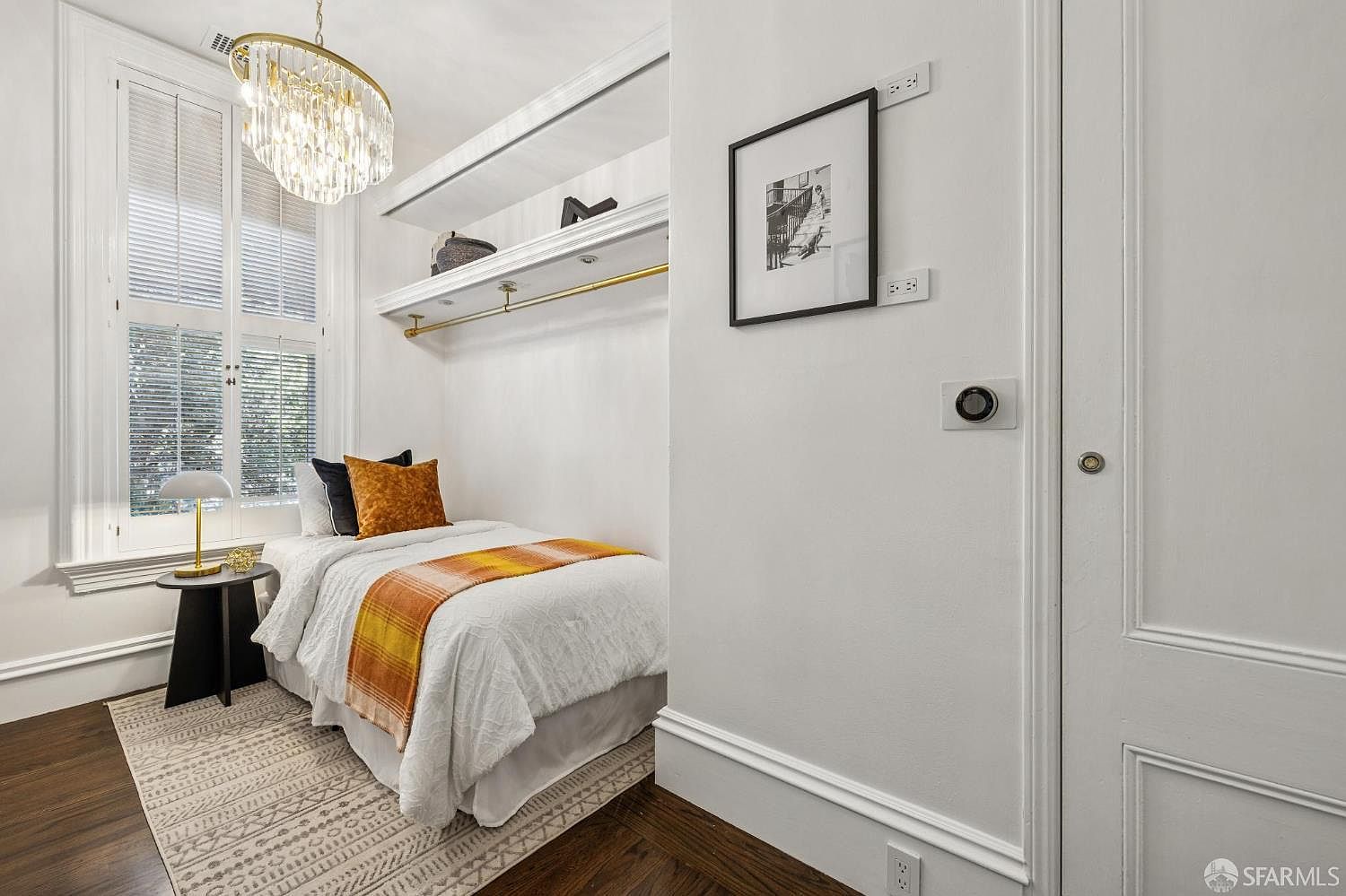 This cozy, compact bedroom features a single bed dressed in white linens with a vibrant orange throw blanket and matching accent pillows. The room is illuminated by a decorative crystal chandelier and a small bedside lamp, while a built-in shelf and hanging rod provide efficient storage. A large window with white shutters allows natural light to brighten the space, which is finished with dark wood flooring and a patterned area rug.