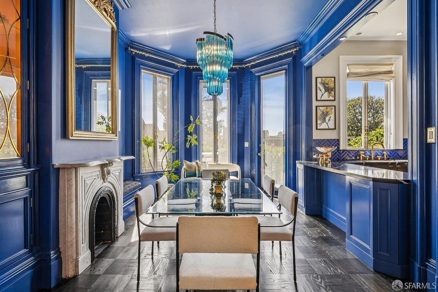 This sophisticated dining room features bold, deep blue walls and trim, creating a dramatic and elegant atmosphere. A glass-topped dining table is centered beneath a striking blue tiered chandelier, flanked by light-toned upholstered chairs. The space includes a classic fireplace with a detailed stone surround, large windows that flood the room with natural light, and a built-in serving area with blue cabinetry and patterned backsplash tiles.
