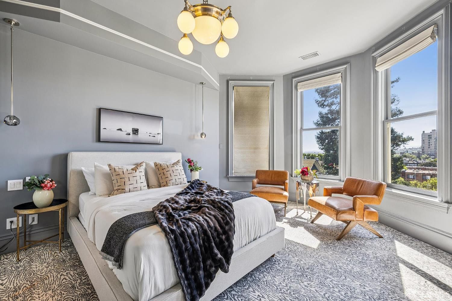 This elegant primary bedroom features a light-toned upholstered bed frame topped with a textured throw blanket and patterned pillows. The room is bathed in natural light from large windows that offer a view of the surrounding neighborhood, complemented by a sophisticated chandelier and modern pendant lighting. Two cognac-colored leather armchairs create a cozy seating area, while the patterned area rug adds depth to the neutral color palette.