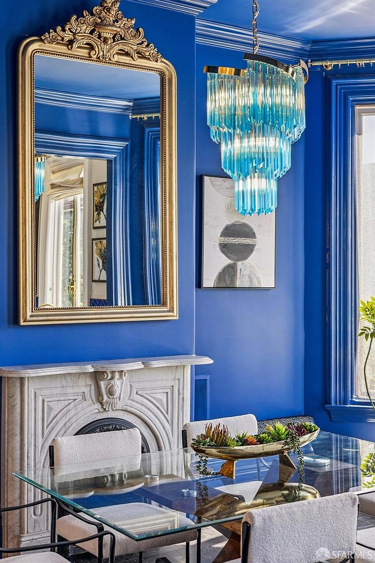 This vibrant dining room features bold blue walls and a striking, tiered blue glass chandelier that serves as a focal point. A glass-topped dining table with light-colored upholstered chairs sits before a classic stone fireplace mantel, which is topped by a large, ornate gold-framed mirror. The room is accented by a modern abstract art piece and a decorative bowl filled with succulents, creating a sophisticated and eclectic atmosphere.