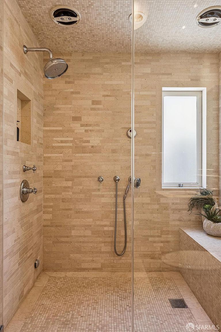 This walk-in shower features textured, light-toned stone tile walls and a matching mosaic tile floor. The space is enclosed by a glass panel and includes a wall-mounted shower head, a handheld sprayer, and a built-in recessed niche for storage. A frosted window provides natural light, while a small bench area adds functionality to the shower enclosure.