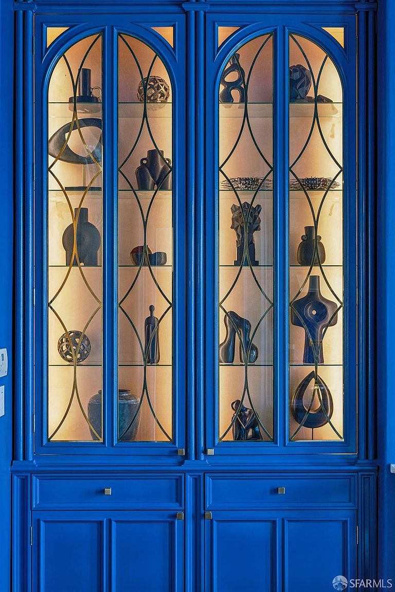 This image showcases a pair of vibrant blue built-in display cabinets featuring elegant, arched glass doors with intricate metal lattice detailing. The interior shelves are illuminated by warm lighting, highlighting a curated collection of dark, sculptural art pieces and decorative objects. The cabinetry is finished with lower storage drawers, providing a sophisticated and artistic focal point for a room.
