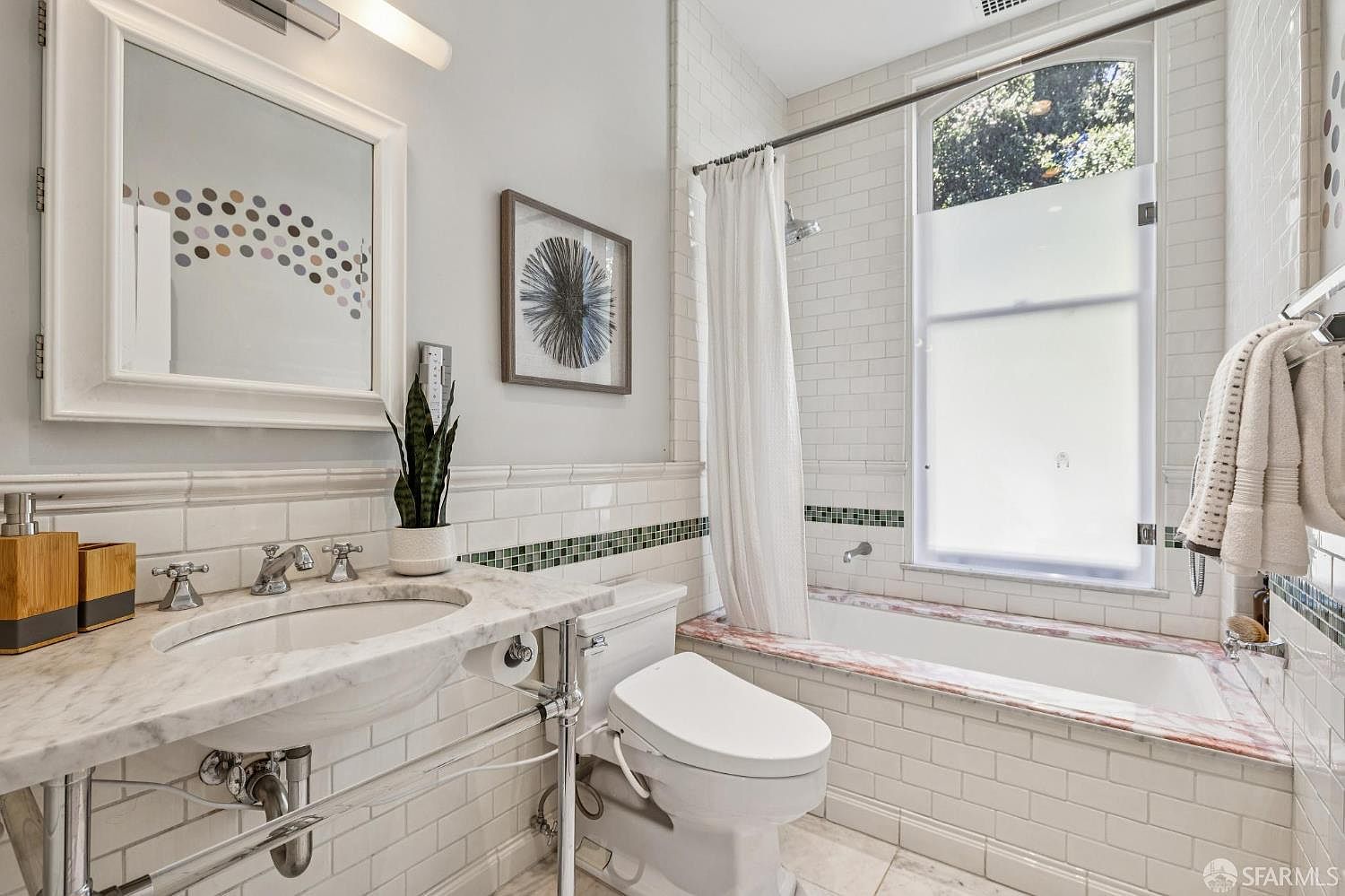 This bright and classic bathroom features white subway tile walls accented by a decorative green mosaic border. A pedestal-style vanity with a stone countertop sits beside a white toilet, while a shower-tub combination is positioned under a large, arched window with frosted privacy glass. The space is well-lit by a wall-mounted vanity light and a framed mirror, creating a clean and timeless aesthetic.
