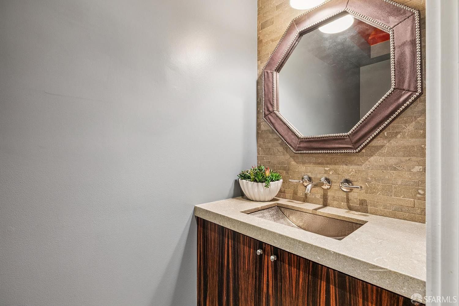 This elegant powder room features a unique hammered metal sink set into a neutral stone countertop atop a dark, vertically-grained wood vanity. The wall behind the vanity is finished with textured, horizontal stone tiles, complemented by an octagonal mirror with a decorative studded frame. A small potted plant adds a touch of greenery to the sophisticated, modern space.