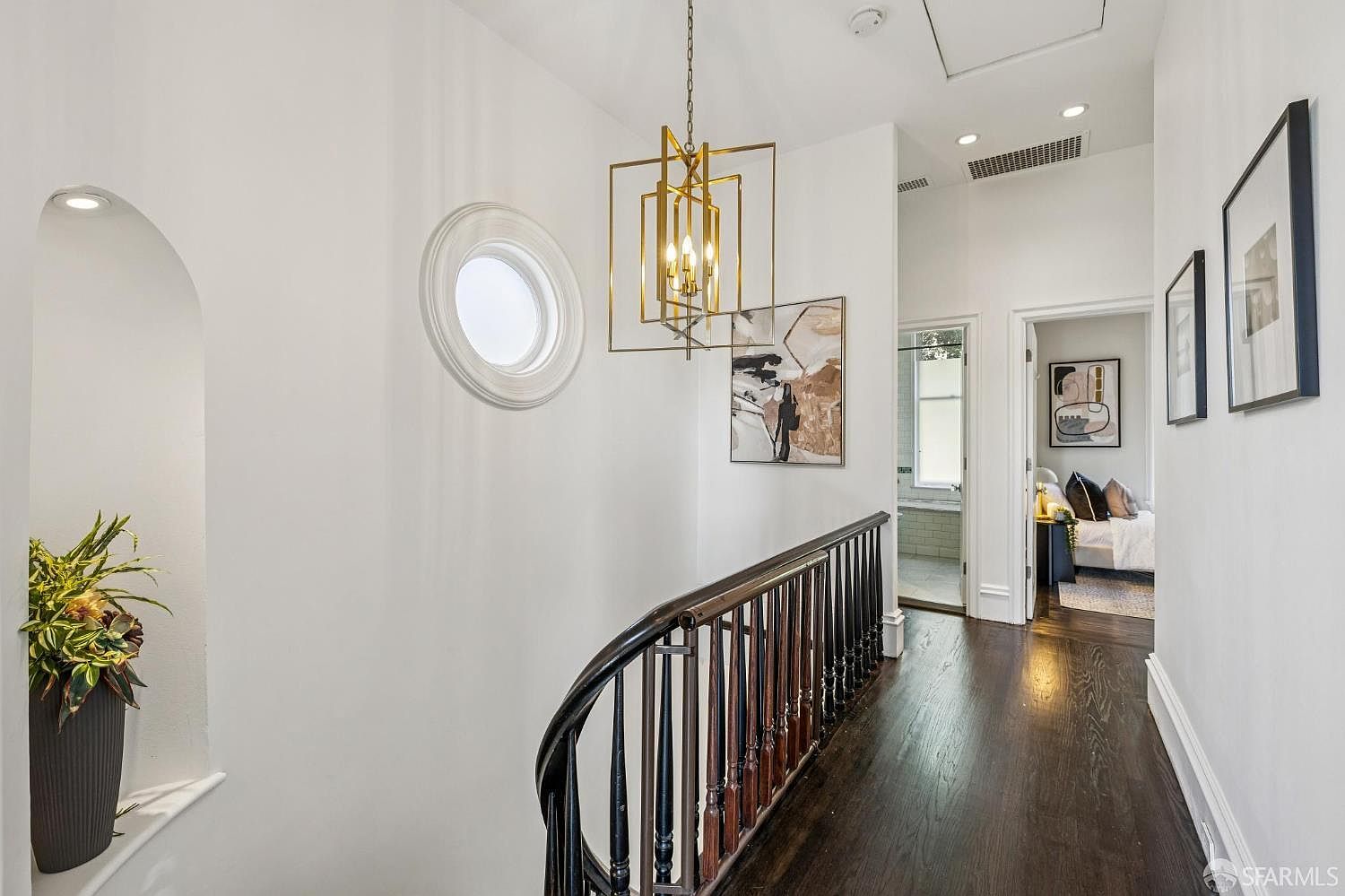 This elegant upper-level hallway features dark hardwood flooring and a curved staircase with a classic dark wood railing and balusters. A striking gold geometric chandelier hangs from the ceiling, complemented by a circular accent window and modern artwork on the walls. The perspective looks down the corridor, showcasing a bright, airy transition space that leads toward a bedroom and bathroom.