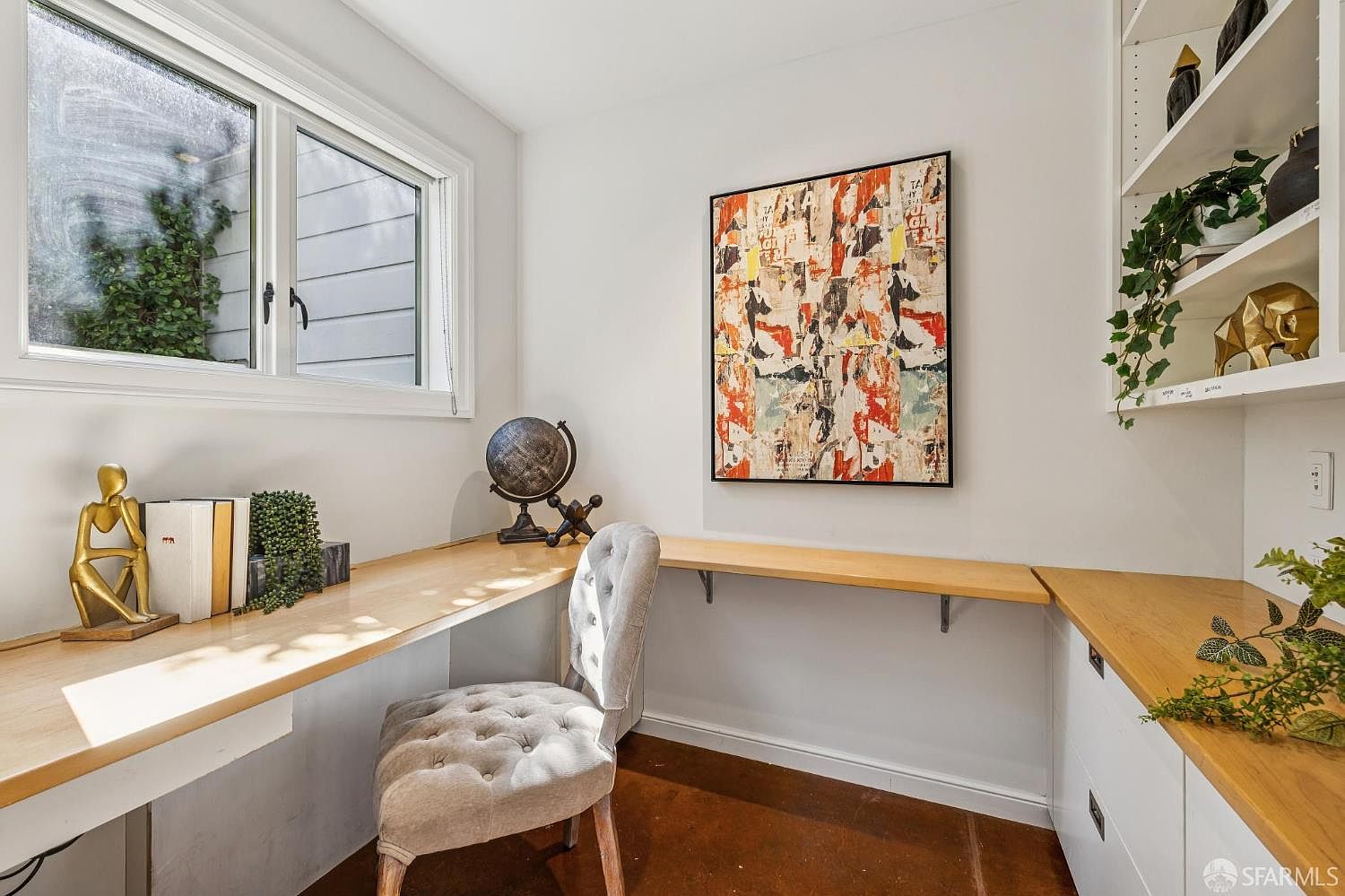 This bright home office features a built-in light wood desk that wraps around the corner, providing ample workspace. A tufted grey chair sits at the desk, which is accented by a globe, books, and decorative sculptures, while a vibrant abstract painting hangs on the wall above. To the right, built-in shelving displays greenery and a gold elephant figurine, creating a functional and stylish workspace.