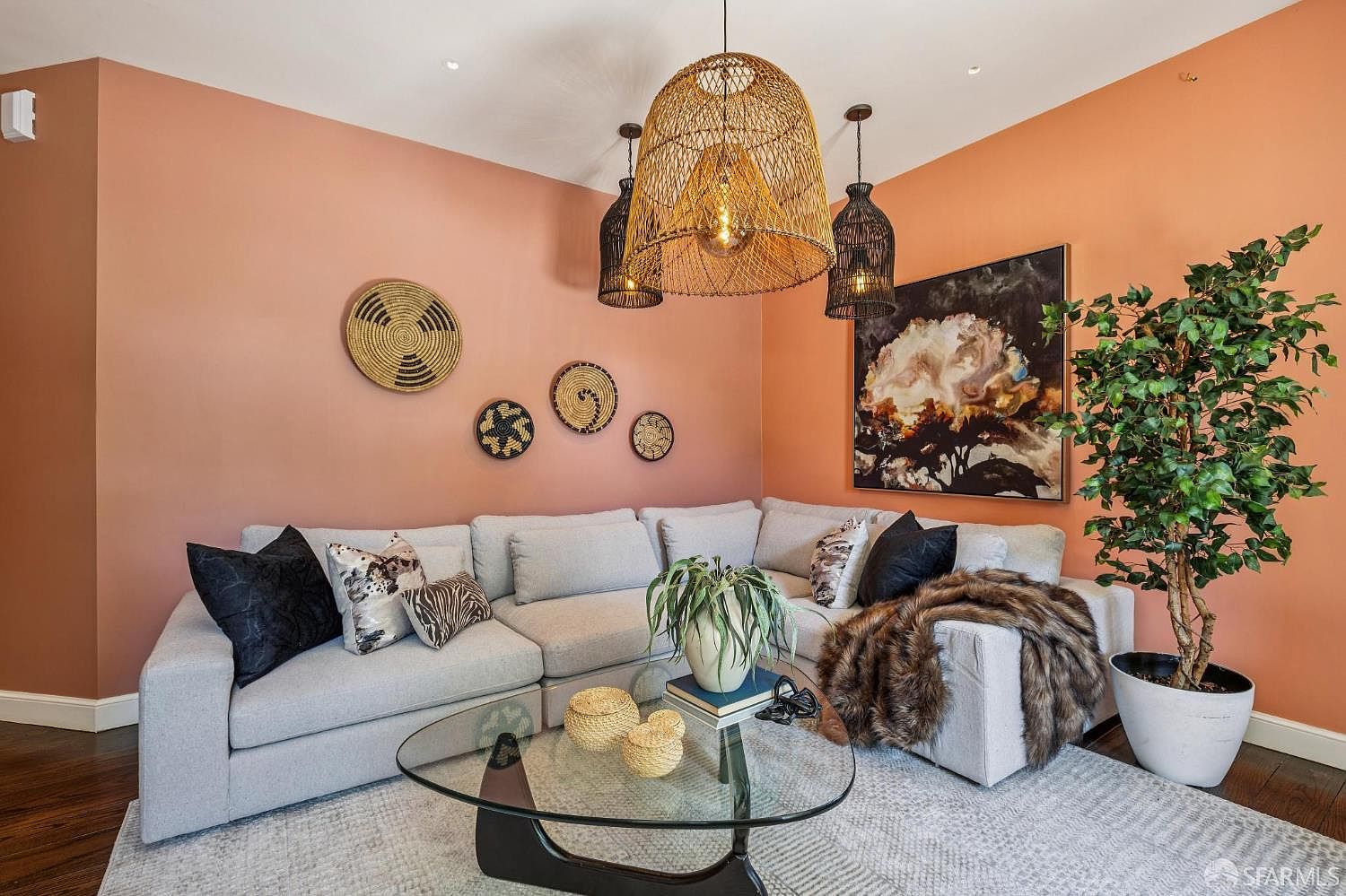 This inviting living room features a large, light-colored sectional sofa arranged against a warm, peach-toned accent wall. The space is decorated with a collection of woven wall baskets, a large abstract art piece, and a striking trio of pendant lights, including a prominent central woven fixture. A glass-topped coffee table sits on a textured area rug, while a potted tree adds a touch of greenery to the corner of the room.
