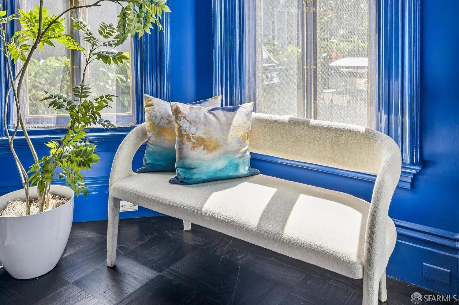 This bright, vibrant space features a cream-colored upholstered bench accented with two decorative pillows featuring abstract blue and gold patterns. The room is defined by bold, deep blue walls and dark, herringbone-patterned wood flooring, creating a striking contrast. Large windows allow natural light to flood the area, highlighting a potted plant in the corner and providing a serene, stylish nook.
