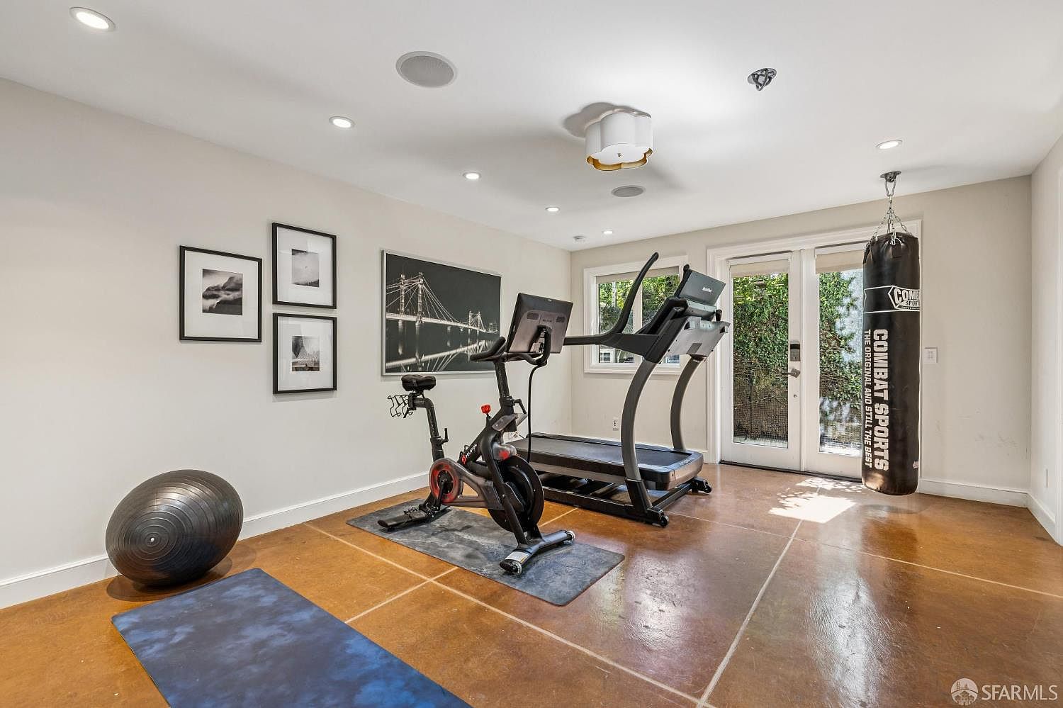 This dedicated home gym features a neutral color palette and durable, large-format tile flooring. The space is equipped with a stationary exercise bike, a treadmill, and a heavy punching bag, making it a functional workout area. A set of glass-paned doors provides natural light and access to the outdoors, while framed artwork adds a personal touch to the walls.