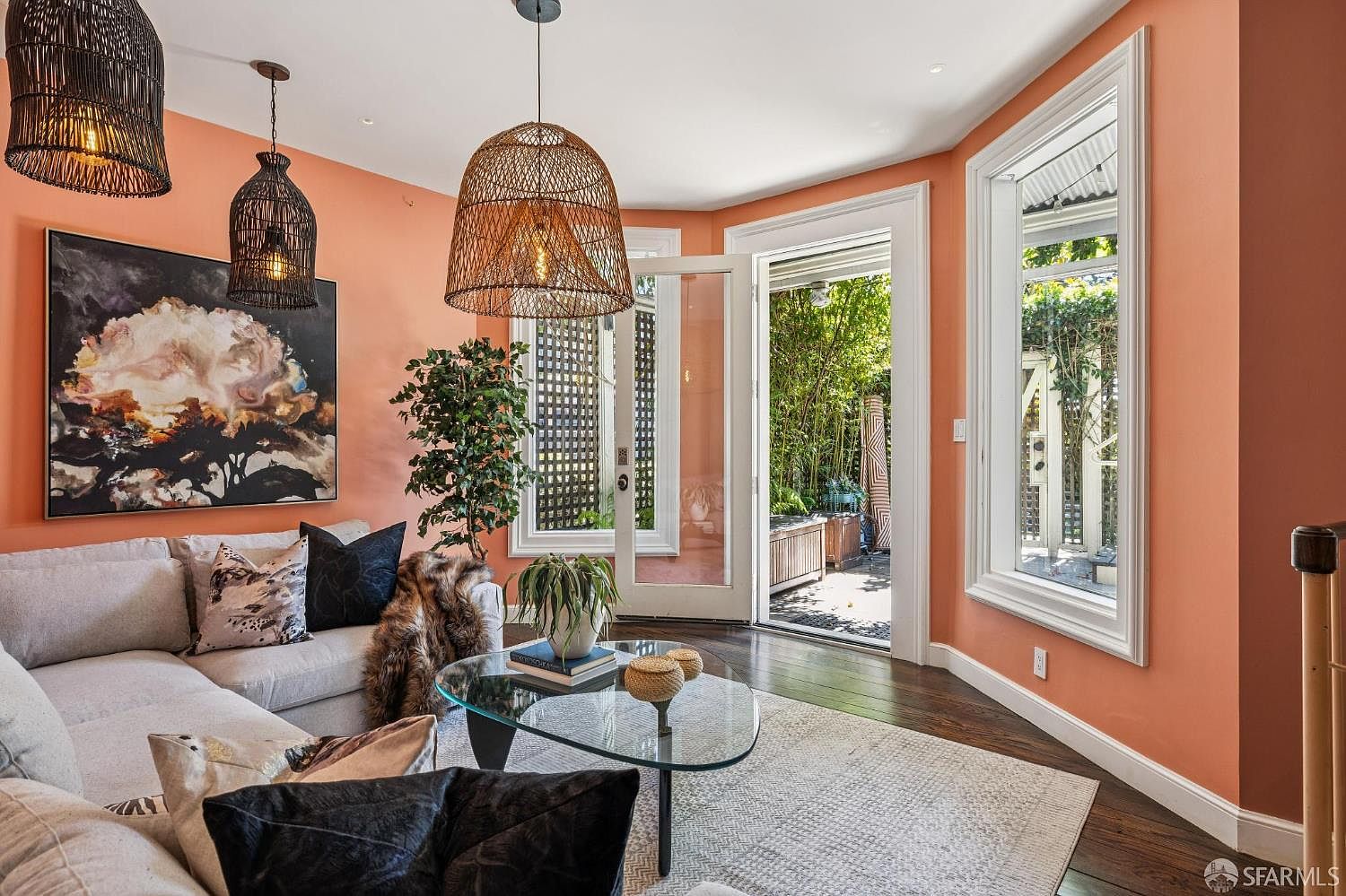 This inviting living room features warm, peach-toned walls and dark hardwood flooring, creating a cozy and vibrant atmosphere. The space is furnished with a comfortable light-colored sectional sofa, a glass-topped coffee table, and distinctive woven pendant lights that add a touch of texture. Large glass doors open directly to a lush, green outdoor garden area, seamlessly blending indoor and outdoor living spaces.