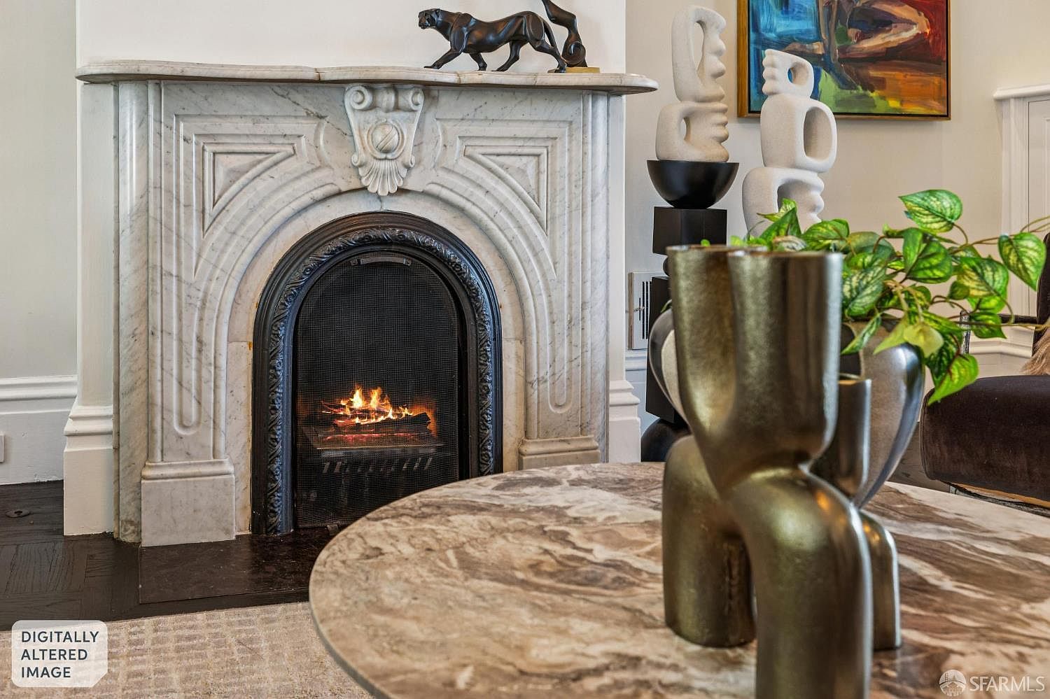 This elegant living room features a prominent stone fireplace with an ornate, carved surround and a dark metal firebox containing a glowing fire. In the foreground, a round stone-topped coffee table holds a sculptural metallic vase and a lush green plant, while abstract white sculptures and a vibrant piece of wall art add a sophisticated, artistic touch. The perspective is captured from a low angle, emphasizing the textures of the fireplace and the curated decor within the room.