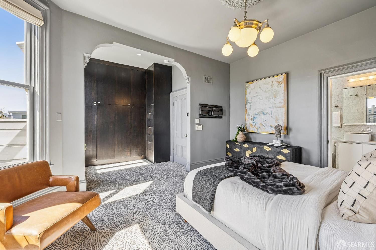 This primary bedroom features a sophisticated design with a large bed topped with a dark throw blanket and patterned pillows. A dark wood wardrobe unit is built into an arched alcove, while a modern dresser sits beneath a large piece of abstract art. The room is illuminated by a decorative chandelier and natural light streaming from a large window, creating an elegant and serene atmosphere.