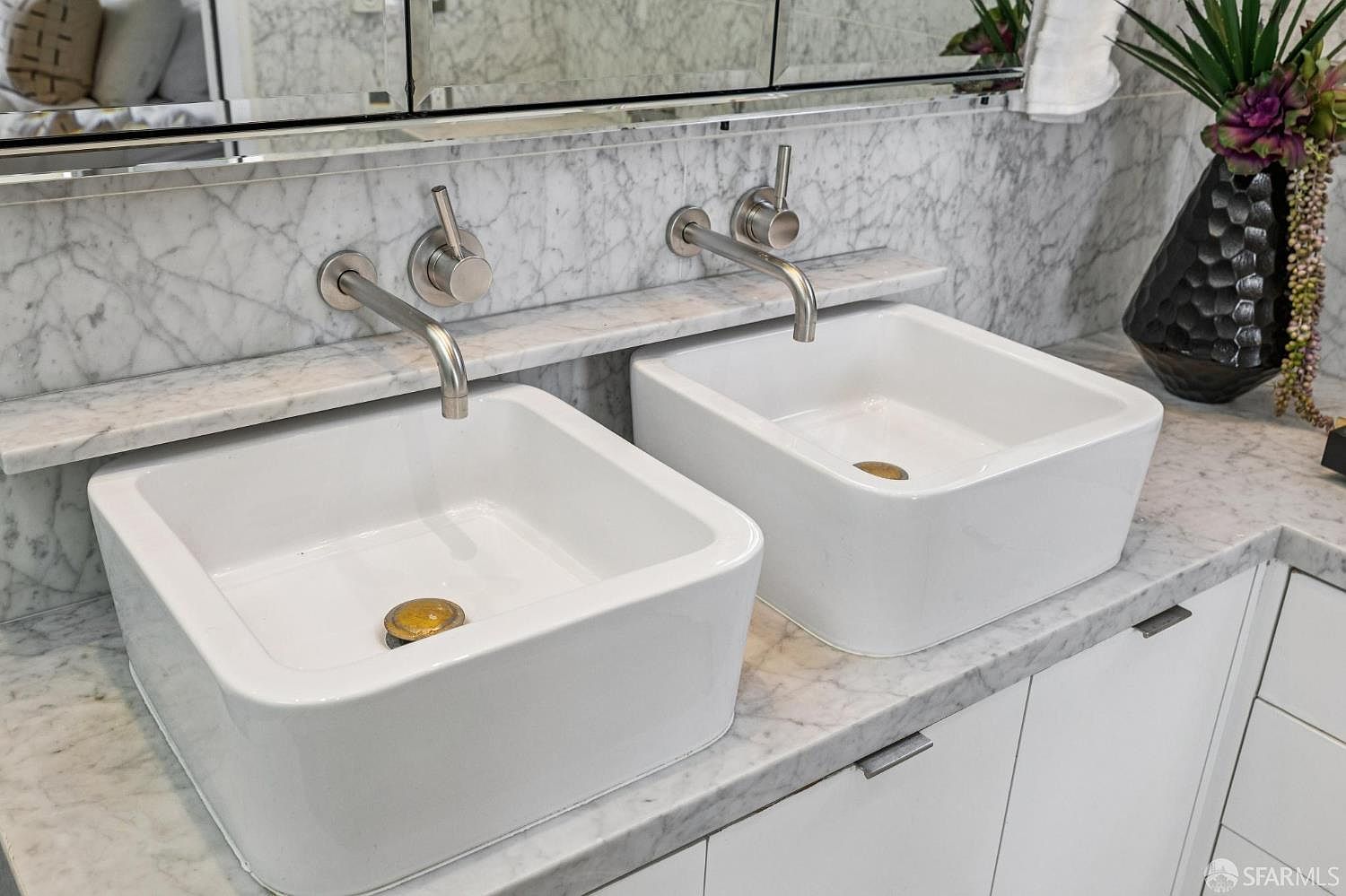 This high-angle shot captures a double vanity setup featuring two square, white vessel sinks mounted on a light-colored stone countertop with distinctive veining. Wall-mounted, brushed-metal faucets and matching handles are installed directly into the stone backsplash, which extends up to a mirrored medicine cabinet. A decorative black vase with artificial greenery sits on the right side of the counter, adding a touch of contrast to the bright, clean aesthetic.