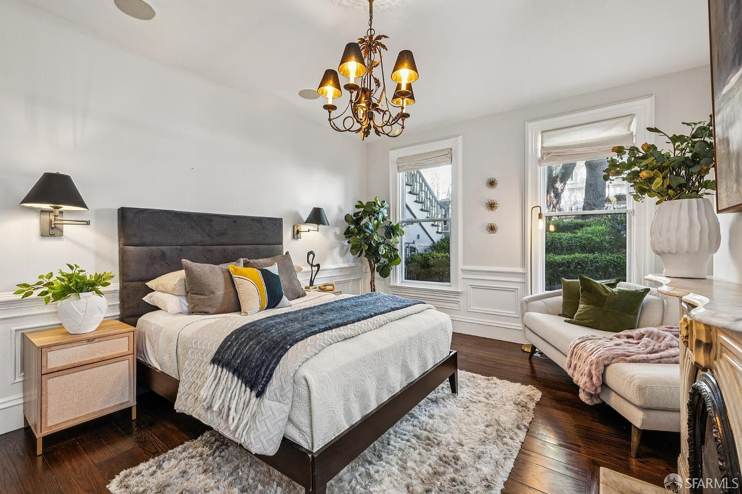This elegant primary bedroom features a plush, dark-toned upholstered headboard and a layered bedding ensemble with a textured throw. The room is anchored by a large, shaggy area rug over dark wood floors and illuminated by a sophisticated chandelier and wall-mounted bedside lamps. A cozy seating area with a light-colored settee and a large potted plant sits near tall windows that provide natural light and a view of the exterior greenery.