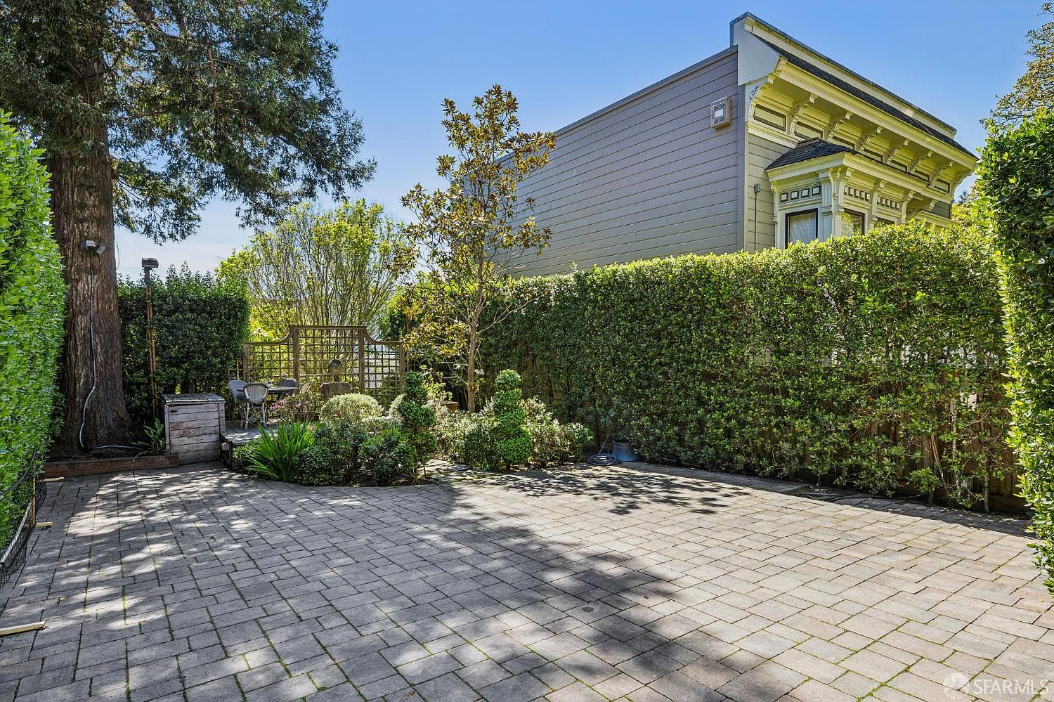 This backyard features a spacious stone-paved patio area that transitions into a lush, private garden enclosed by tall, manicured hedges. A large mature tree stands on the left, while a small wooden deck with outdoor seating provides a cozy nook near the back of the property. The scene is captured from a ground-level perspective, showcasing the home's classic architectural siding and decorative trim against a clear blue sky.