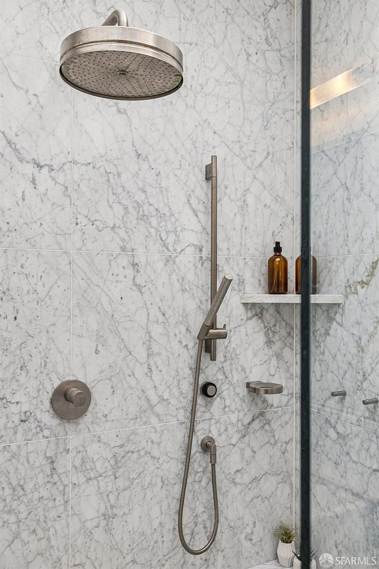 This close-up shot captures a modern shower stall featuring light-colored stone wall tiles with prominent grey veining. The shower is equipped with a large circular rainfall showerhead, a handheld sprayer on a vertical slide bar, and a built-in stone shelf holding amber-colored bottles. A glass partition with a dark frame separates the shower area, creating a sleek and contemporary aesthetic.
