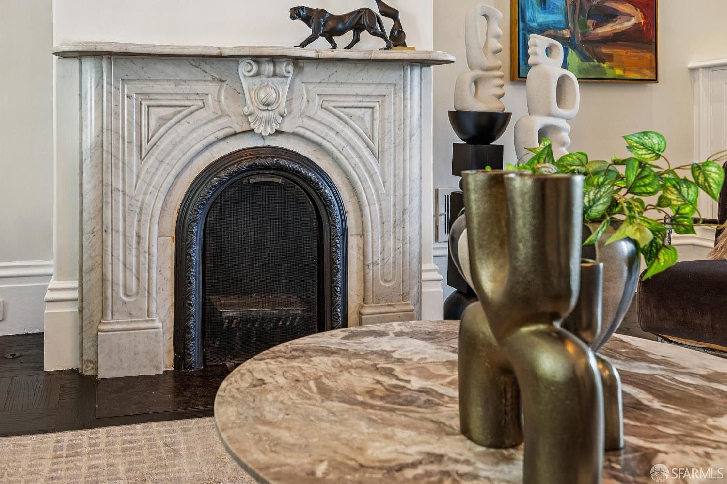 This elegant living room space centers on a classic stone fireplace with an arched firebox and a detailed mantelpiece. A round, stone-topped coffee table sits in the foreground, adorned with a sculptural metallic vase and a lush green plant. The room is decorated with sophisticated art pieces, including a black panther figurine on the mantel and abstract white sculptures, creating a refined and artistic atmosphere.