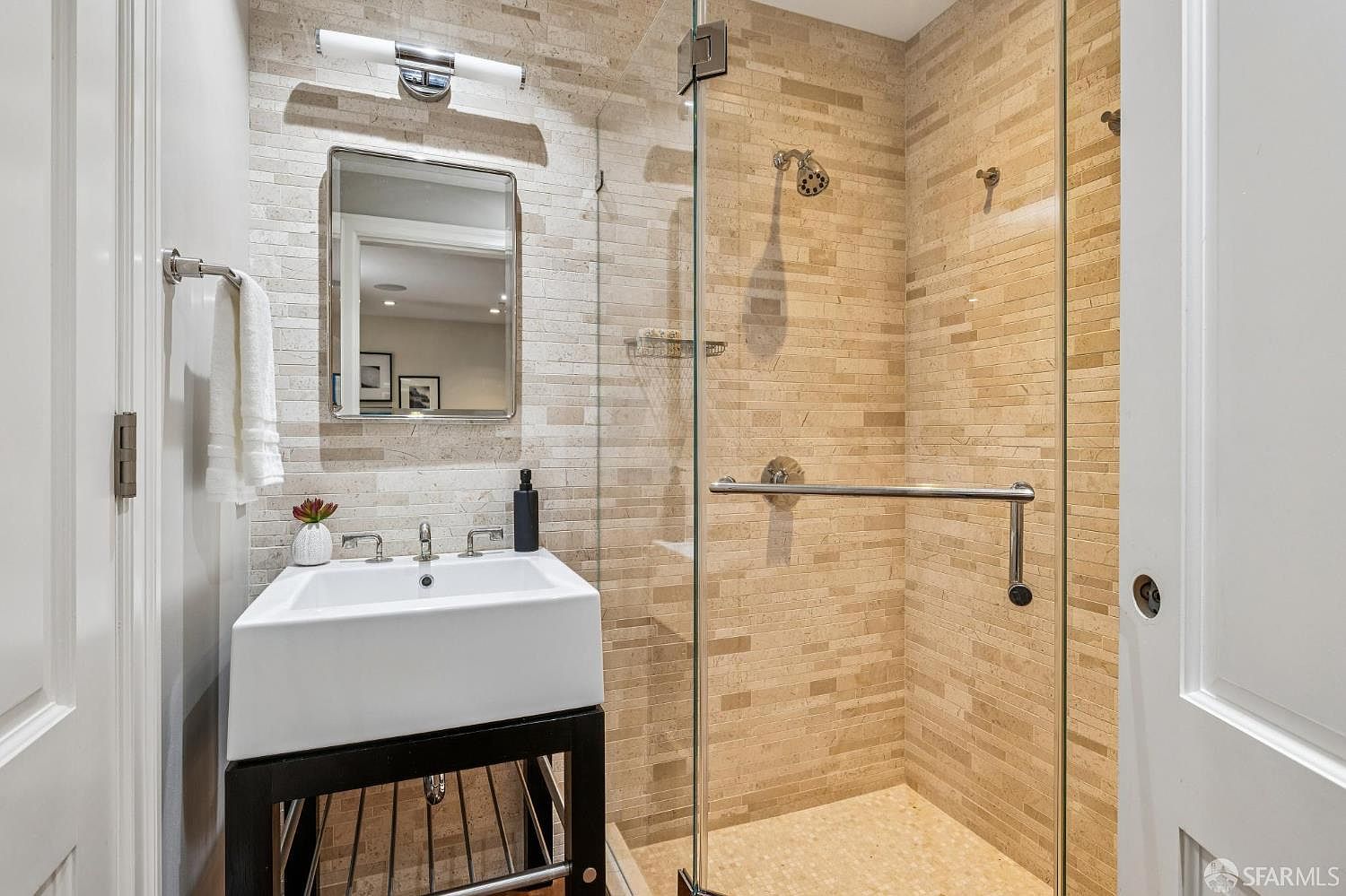 This compact, modern bathroom features a sleek pedestal-style vanity with a white basin and a rectangular mirror mounted on a textured stone-tiled wall. To the right, a glass-enclosed shower stall showcases matching stone tile work and a polished chrome shower head. The space is well-lit by a contemporary wall-mounted light fixture, creating a clean and sophisticated aesthetic.