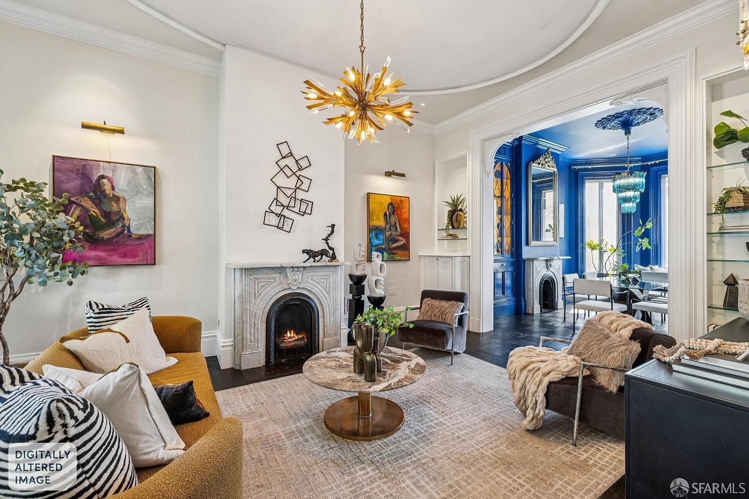 This elegant living room features a sophisticated blend of classic architectural details and modern decor, anchored by a central fireplace with a stone surround. A striking gold starburst chandelier hangs above a circular stone-topped coffee table, while a plush mustard-toned sofa and dark accent chairs provide comfortable seating. The room flows seamlessly into a vibrant, deep blue dining area, creating a dramatic contrast that highlights the home's refined and artistic aesthetic.