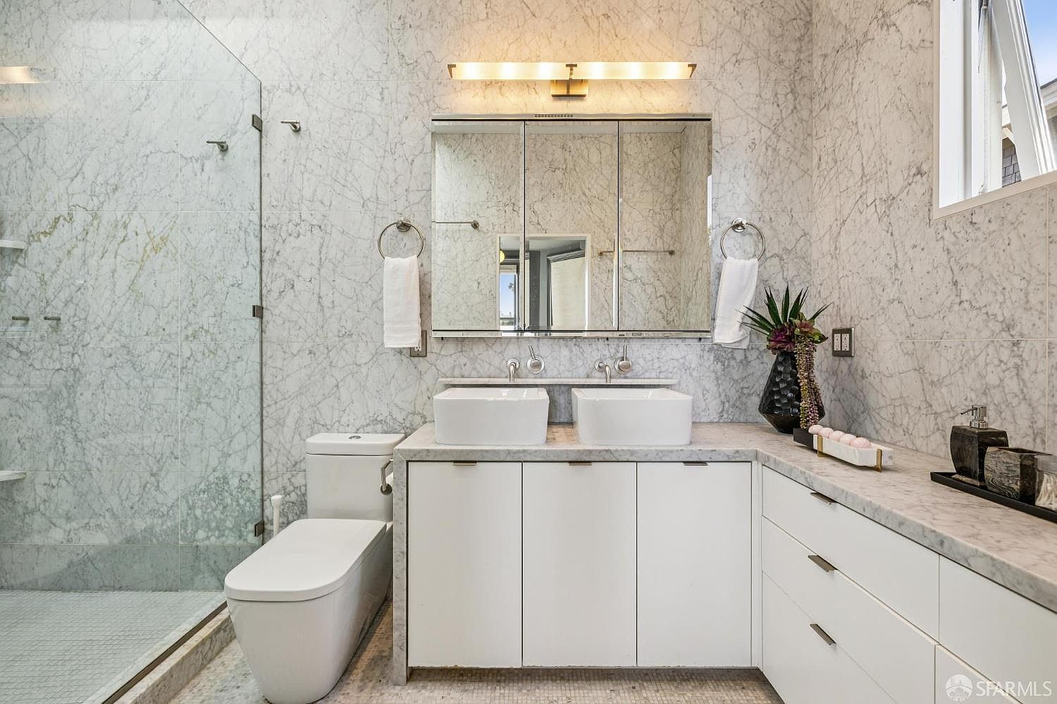 This modern primary bathroom features a double vanity with white cabinetry and a light-toned stone countertop, complemented by a large, multi-paneled medicine cabinet. The walls and shower area are finished in a light-colored, veined stone tile, creating a cohesive and luxurious aesthetic. A glass-enclosed shower and a sleek, wall-mounted toilet complete the space, which is illuminated by a contemporary horizontal light fixture.