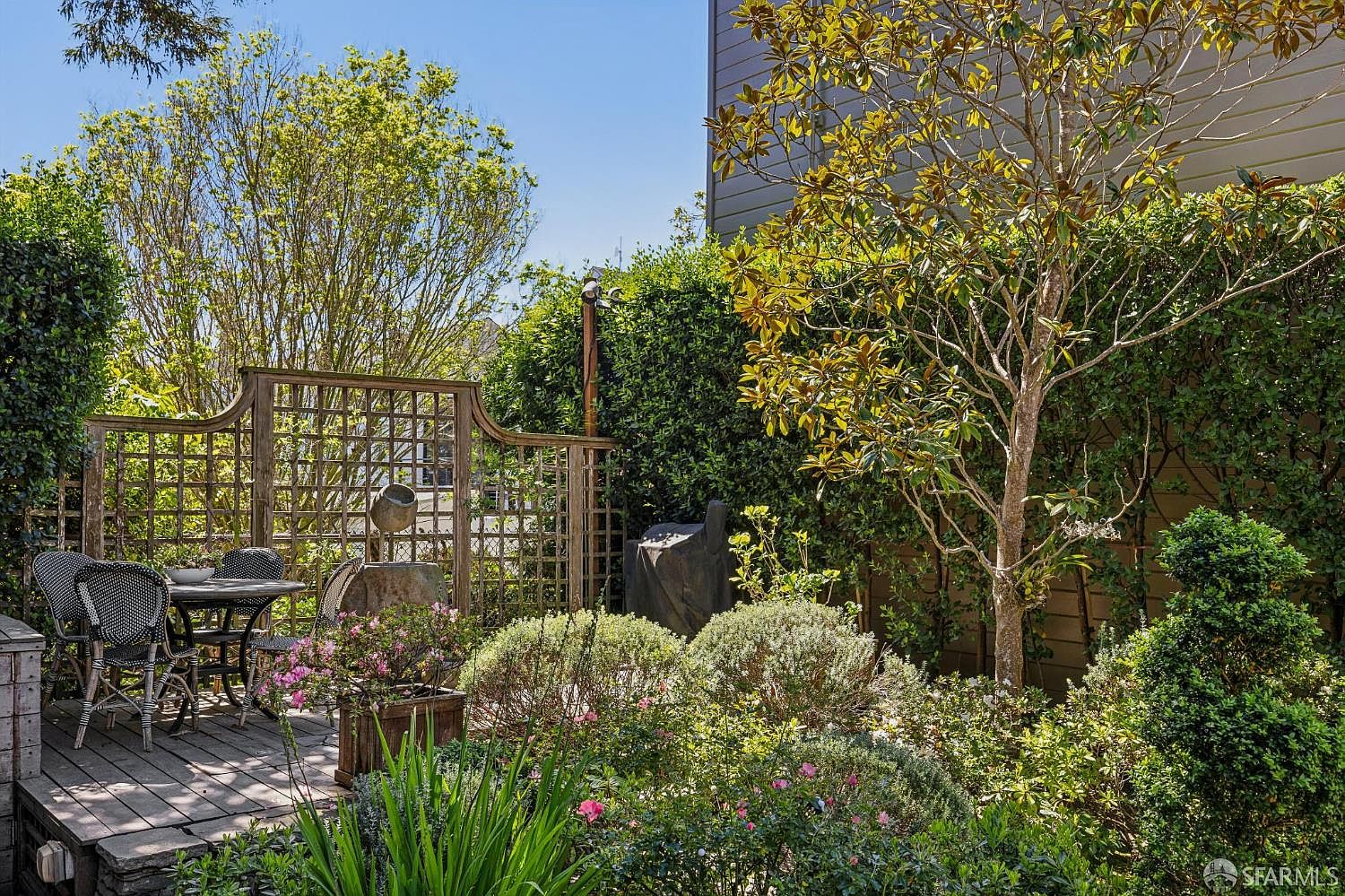 This charming outdoor space features a wooden deck with a bistro-style dining set, perfect for intimate gatherings. The area is framed by a decorative wooden lattice screen and lush, mature landscaping, including a prominent magnolia tree and dense hedges that provide privacy. The scene captures a serene, sun-drenched garden atmosphere with a mix of manicured shrubs and flowering plants.