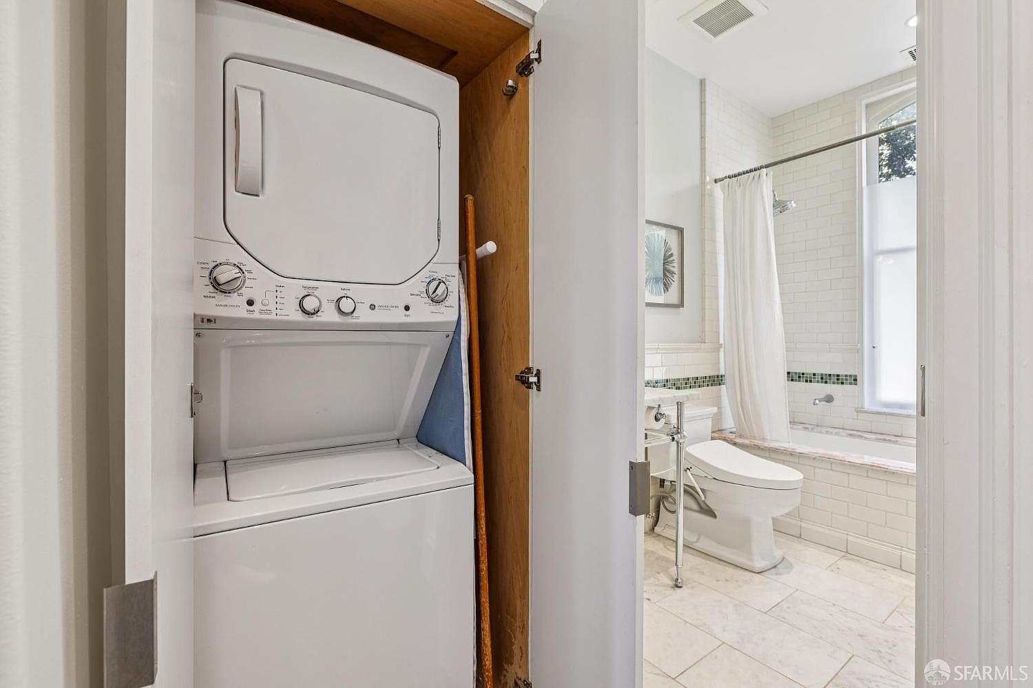 This image captures a compact laundry closet featuring a stacked washer and dryer unit, conveniently tucked away behind a door. Adjacent to the laundry area is a bright, well-lit bathroom with light-colored tile flooring, a white toilet, and a bathtub with a shower curtain. The perspective looks from the laundry closet into the bathroom, highlighting the efficient use of space in this home layout.