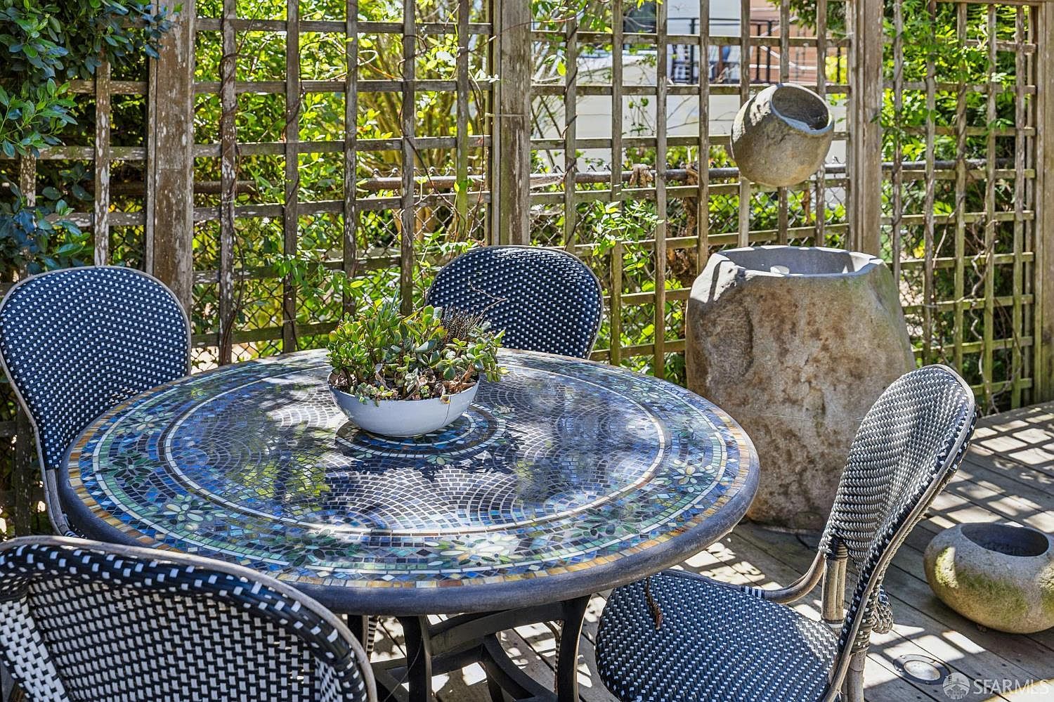This outdoor space features a circular table with a decorative mosaic tile top, surrounded by four woven-patterned chairs on a wooden deck. A stone water feature stands against a wooden lattice fence, creating a serene and private atmosphere. The scene is captured from a slightly elevated perspective, highlighting the intimate dining area and the lush greenery in the background.