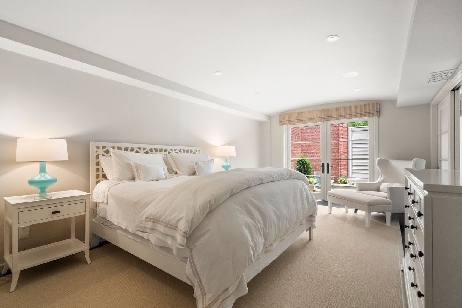 This bright and airy primary bedroom features a white bed frame with a decorative cutout headboard, dressed in crisp white linens. To the right, a matching white dresser and a comfortable armchair with an ottoman create a cozy seating area. Large glass doors lead to an outdoor space, allowing natural light to fill the room, which is finished with neutral carpeting and soft, recessed lighting.